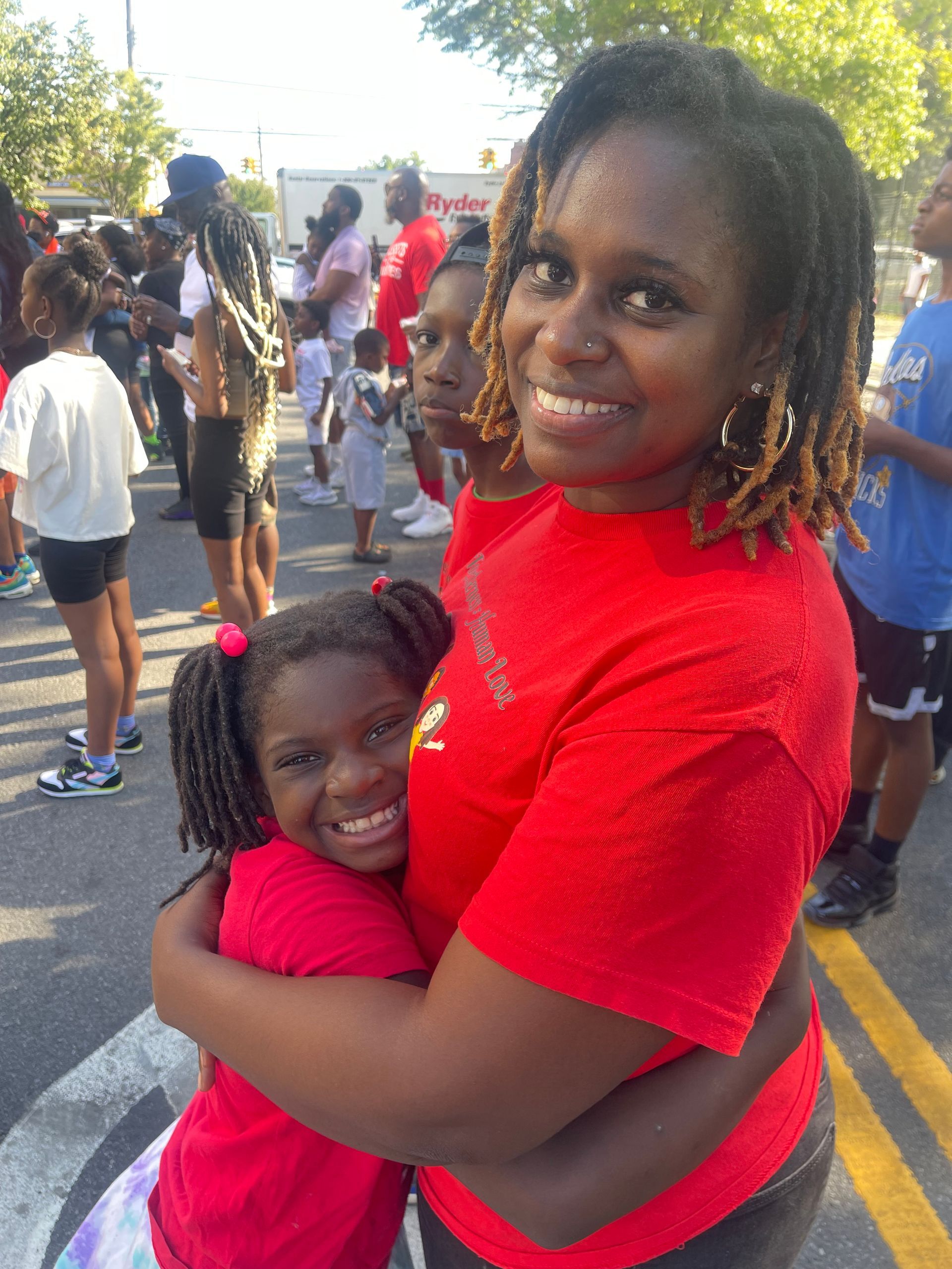Woman in red shirt hugs a girl in a red shirt. Both smile. People in background, outside.