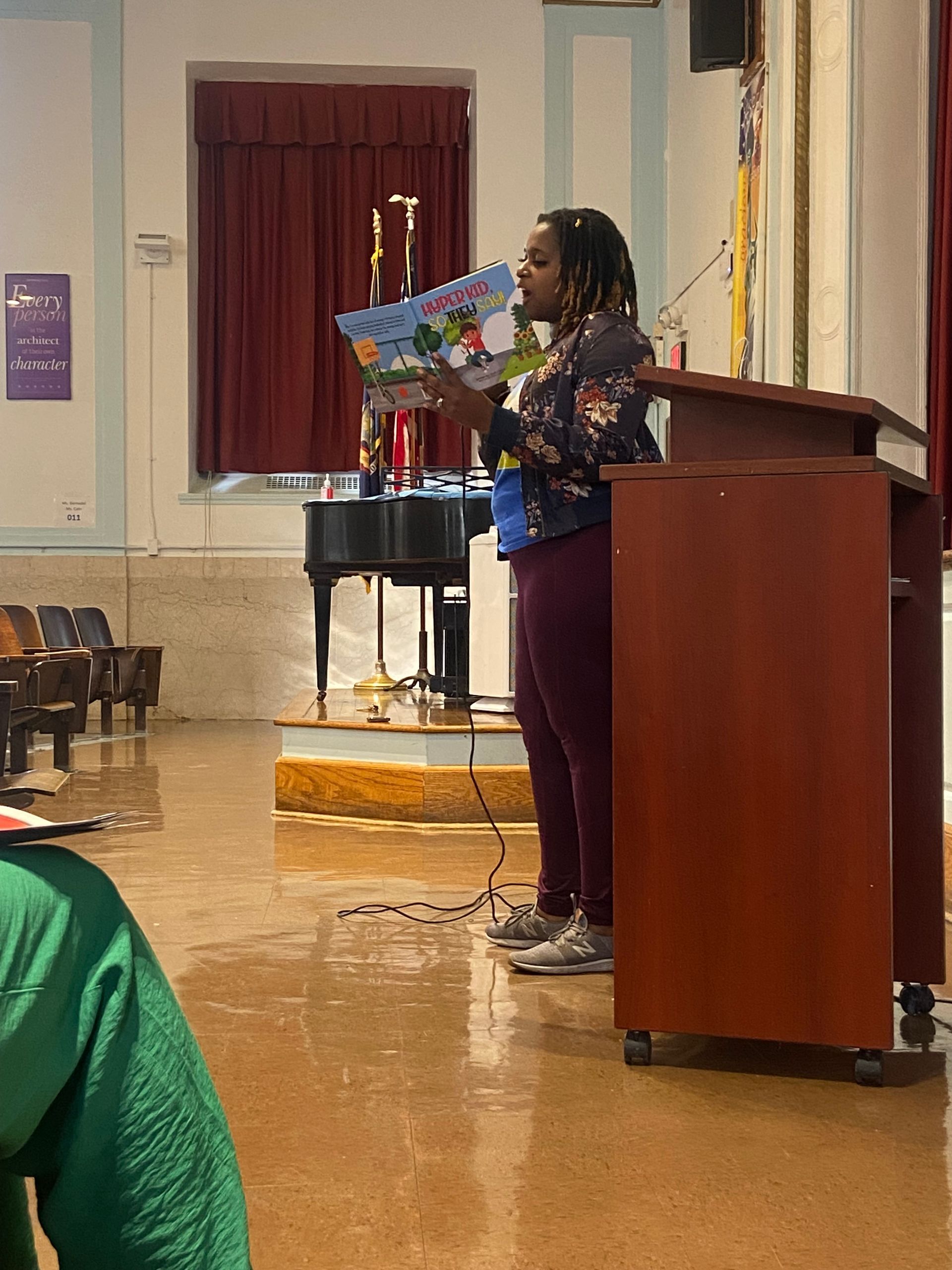 Woman reading a book at a podium in a hall, holding a colorful children's book.
