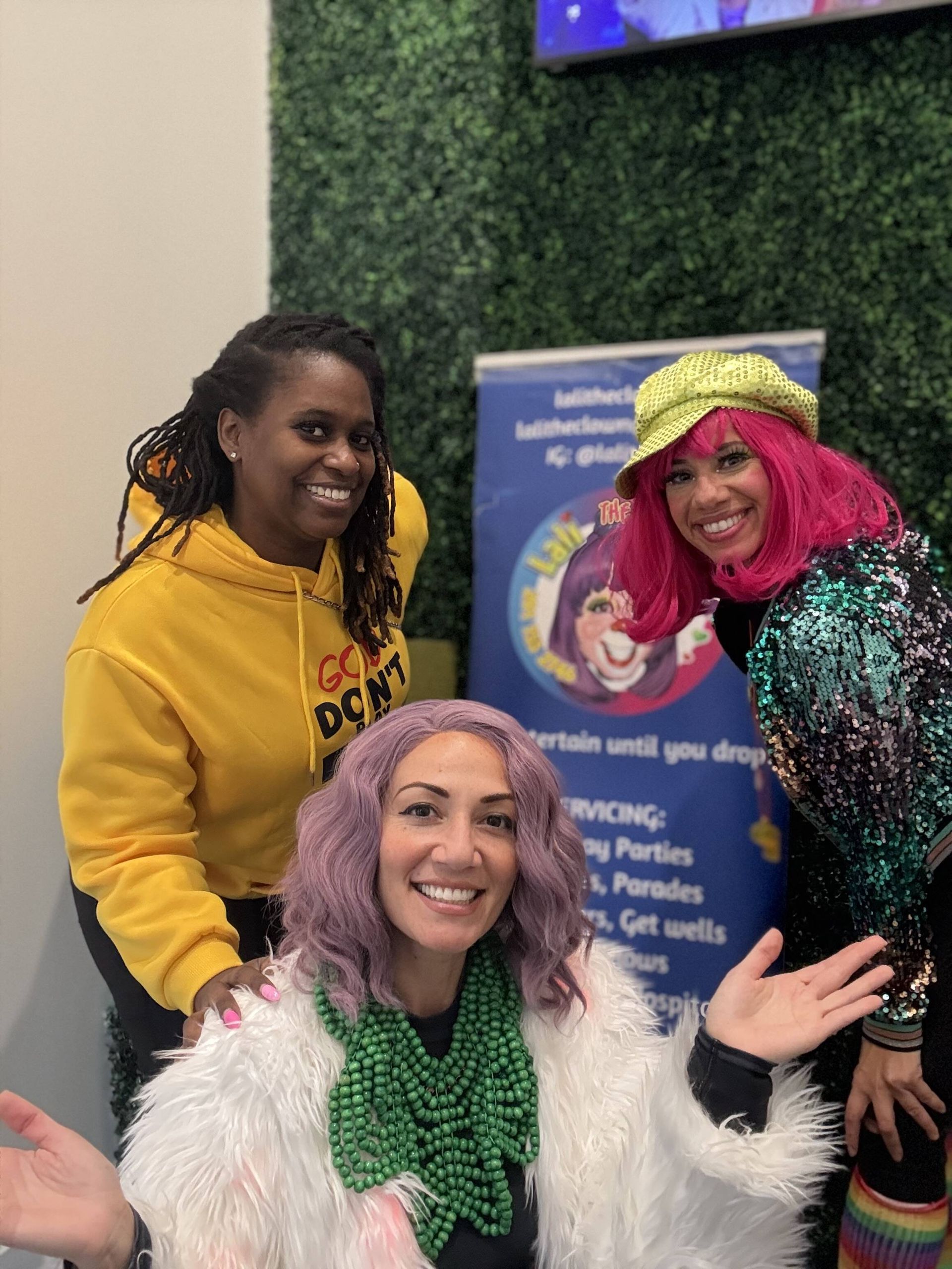 Three people smiling together: A woman in a yellow hoodie, one in a pink wig, and a woman in purple hair and white fur.