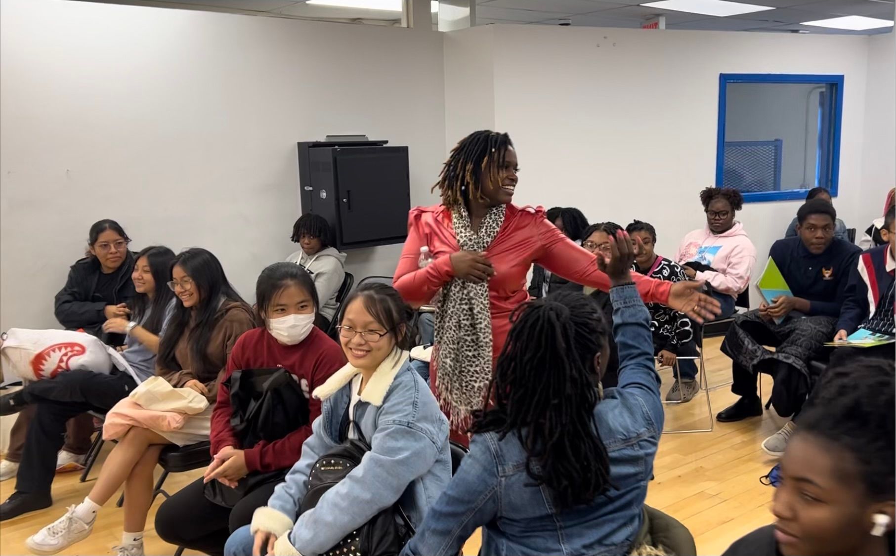 Woman in red dress speaking to a group of seated students in a white-walled room.
