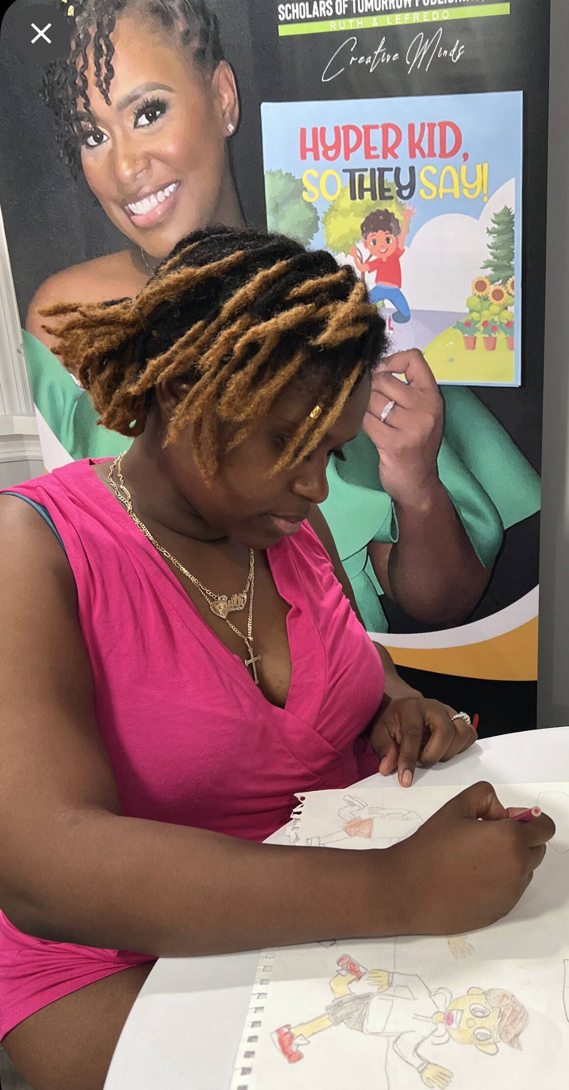 Woman in pink dress signing a book, in front of a poster of herself and a children's book.