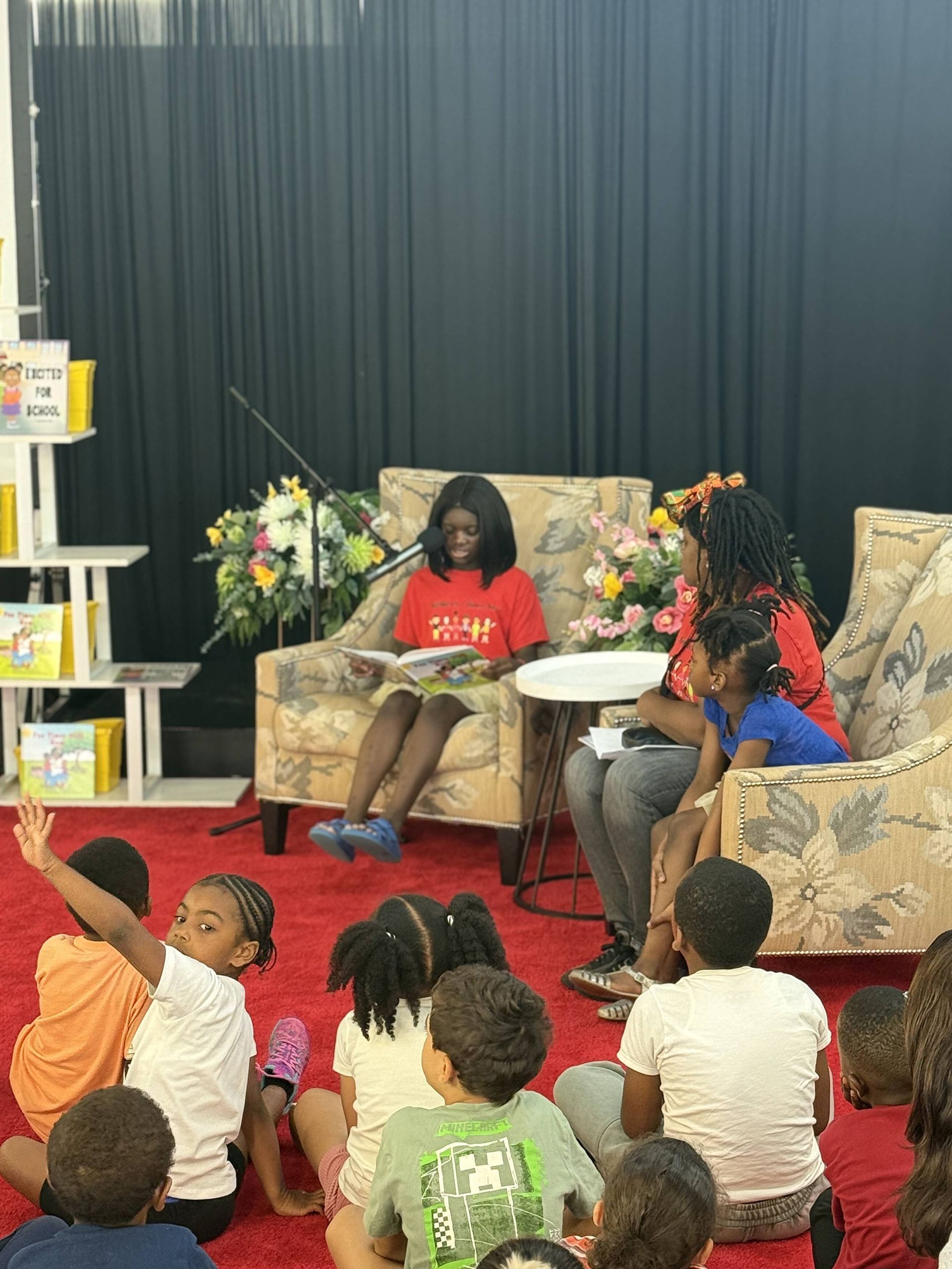 A person reads to children seated on a red carpet. Flowers and books are in the background.