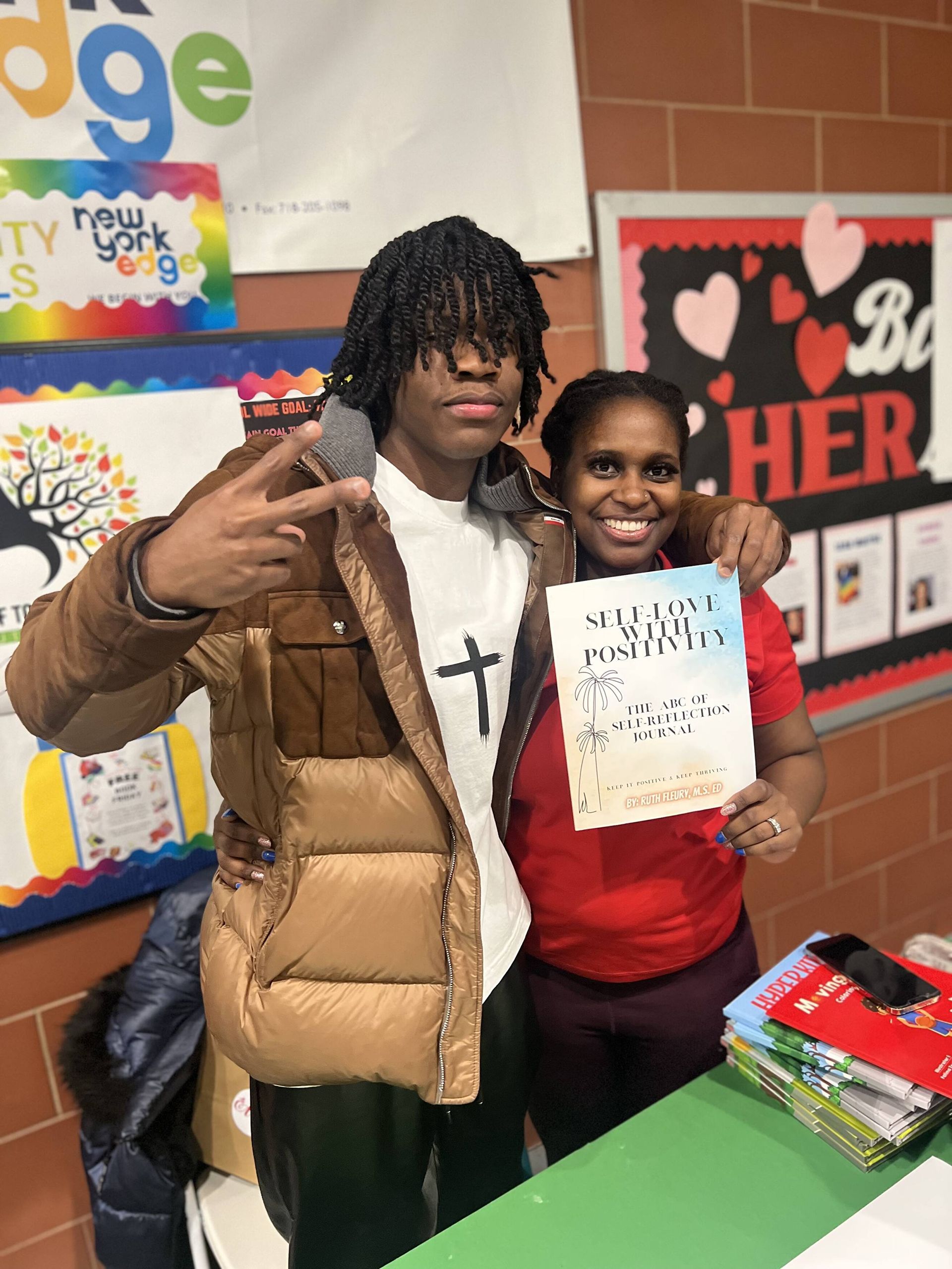 Teenager and woman smiling, holding paper. Teenager gestures peace sign, wearing jacket. Background with school decorations.