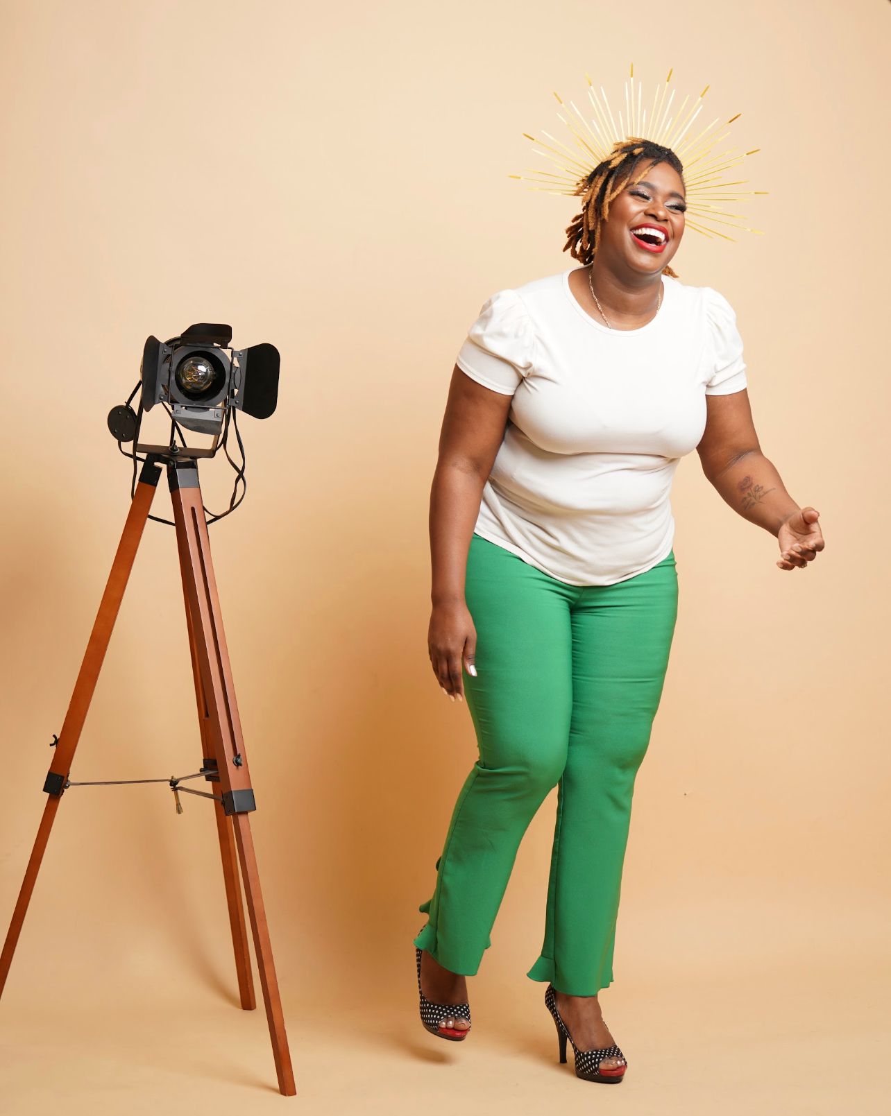 Woman laughing, posed beside camera on tripod, wearing white top and green pants, gold halo, on tan background.