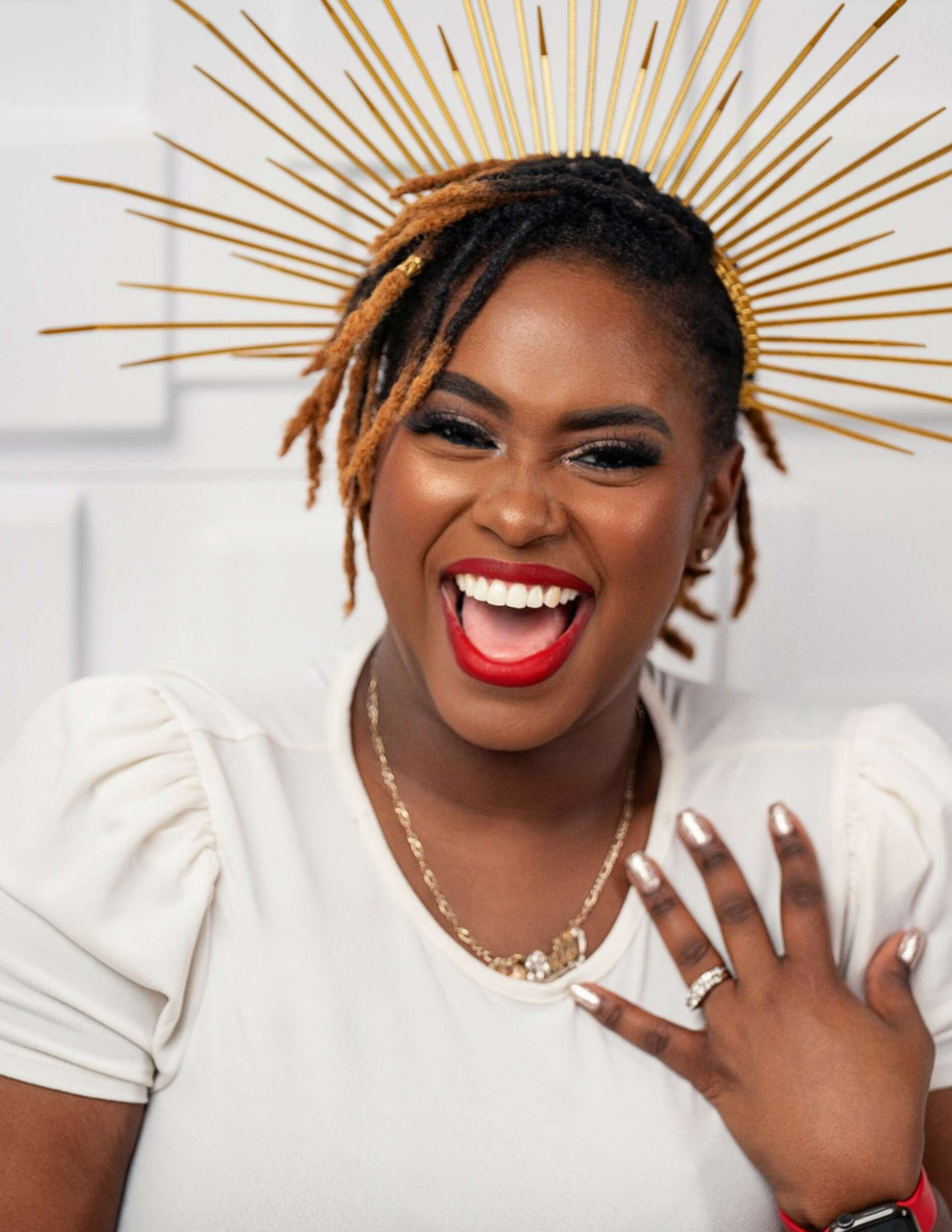 Woman laughing, wearing gold halo, white top, red lipstick, hand on chest, gold necklace.