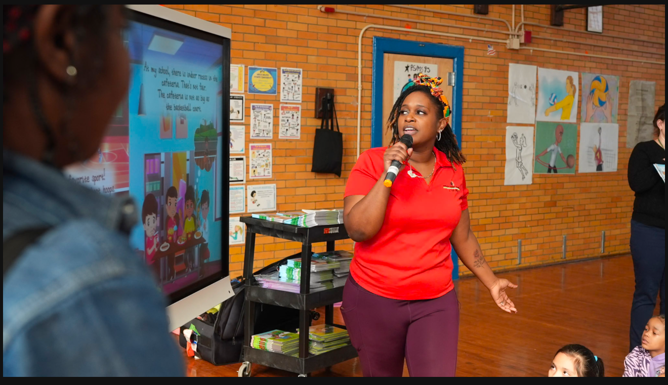 Woman in red shirt speaking into a microphone, standing in front of a screen with a cartoon and a group of people.