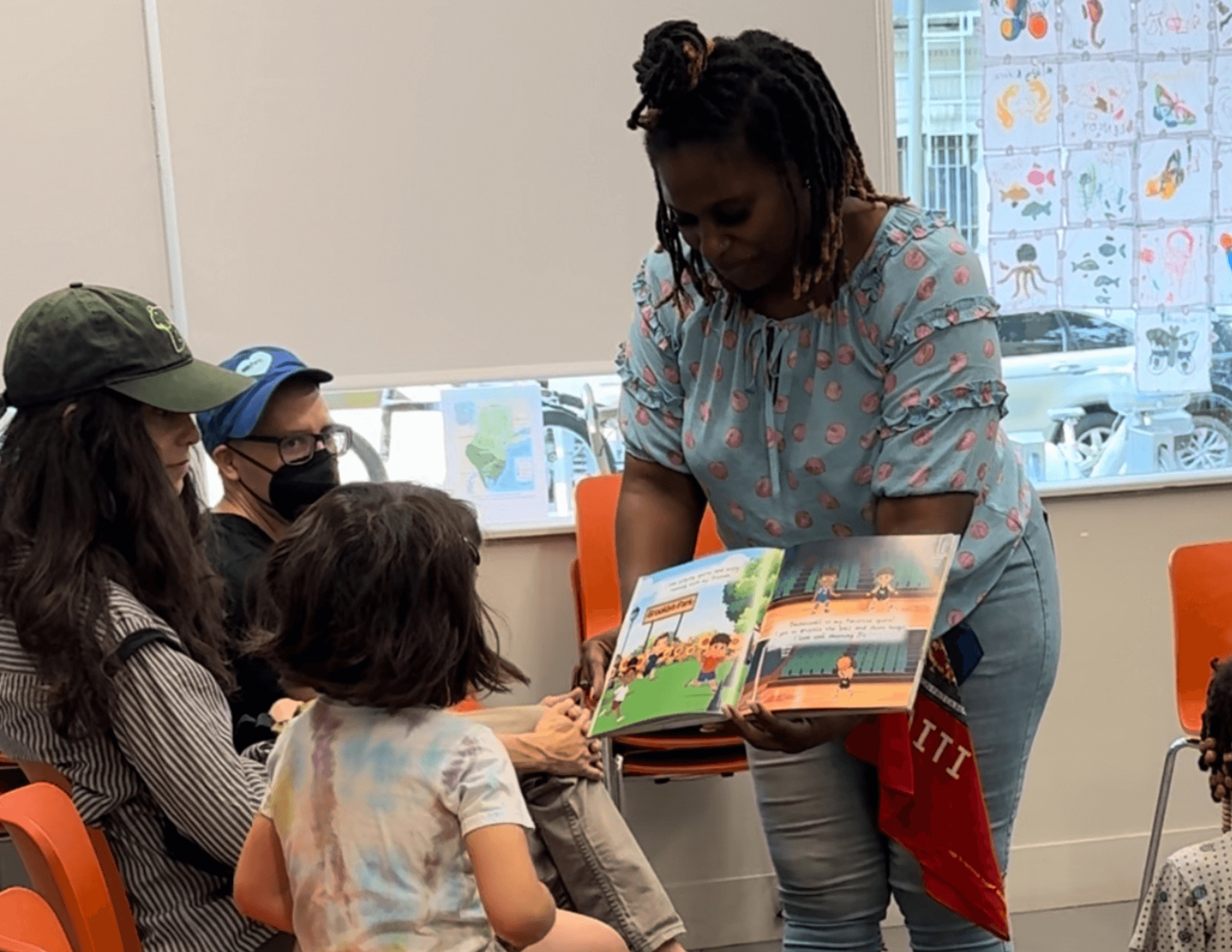 Person reading a book to a group. Others seated and looking on. Indoors, sunny.