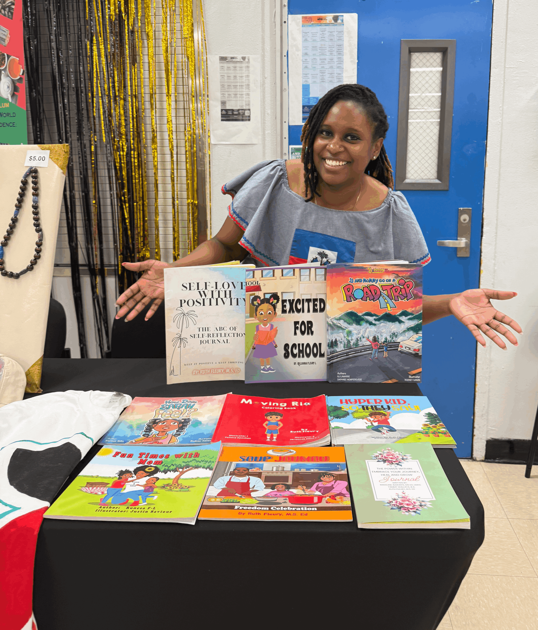 Woman with books, smiling, arms outstretched, behind a table, indoors.