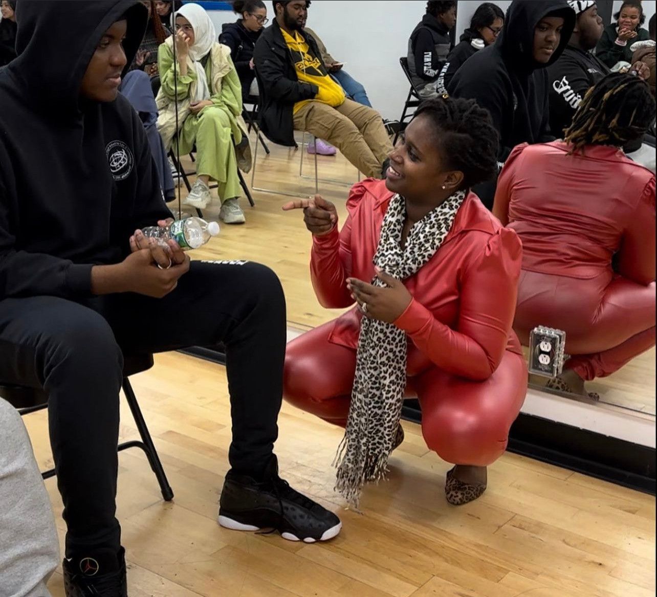 Woman in red outfit squats, points at person in black hoodie; group of people in background.