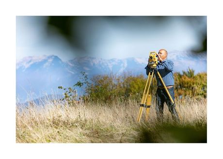 man in jeans looking through a surveying tripod with mountains in the background