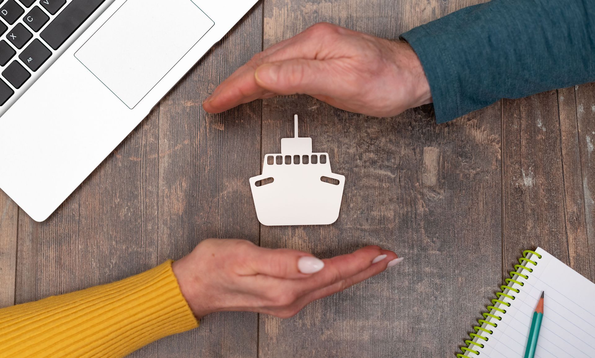 Two hands framing a small ship-shaped cutout on a desk beside a laptop and notebook. Two hands framing a small ship-shaped cutout on a desk beside a laptop and notebook.