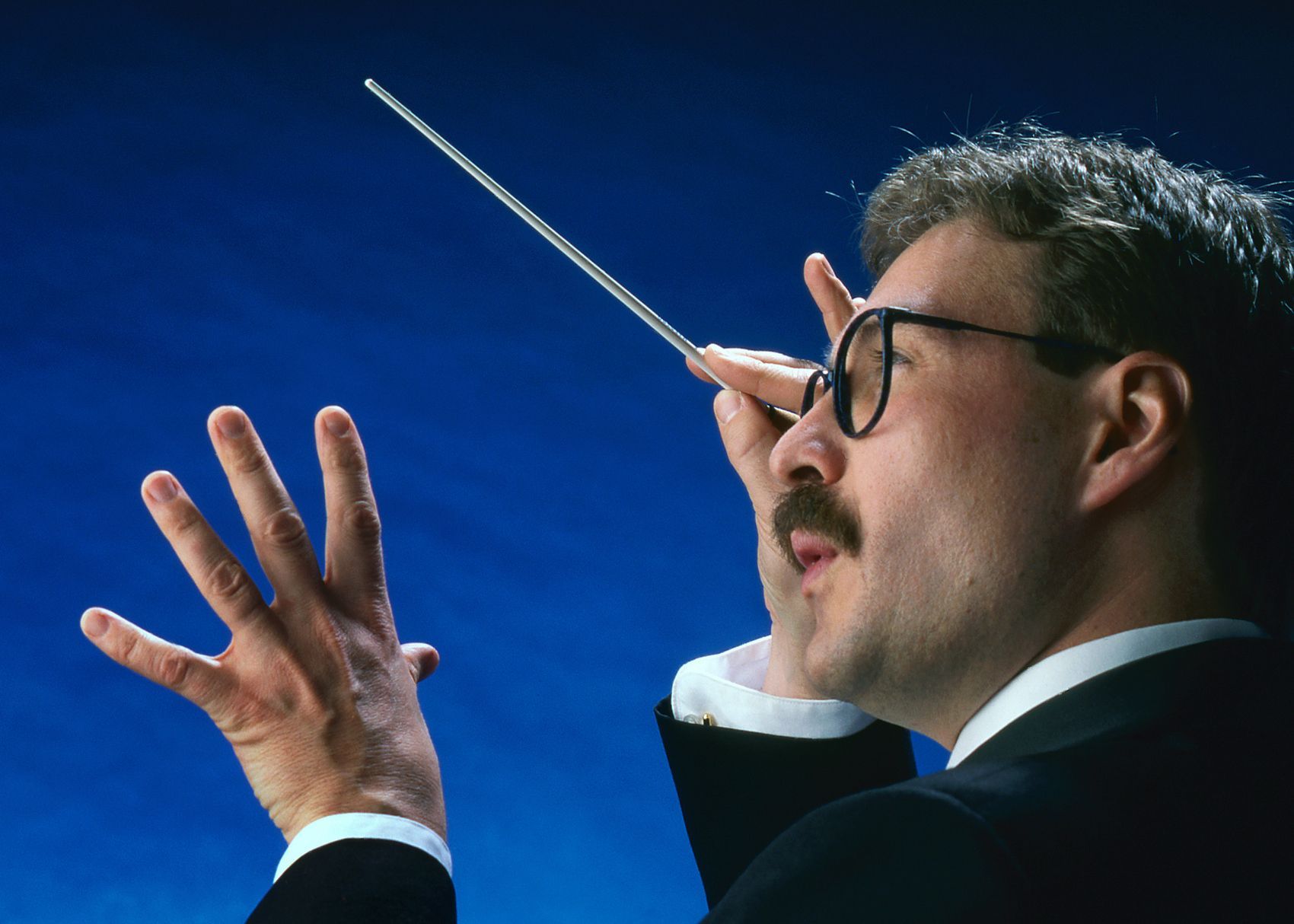 Man in suit conducting an orchestra with a baton against a blue background, left hand open, focused expression.