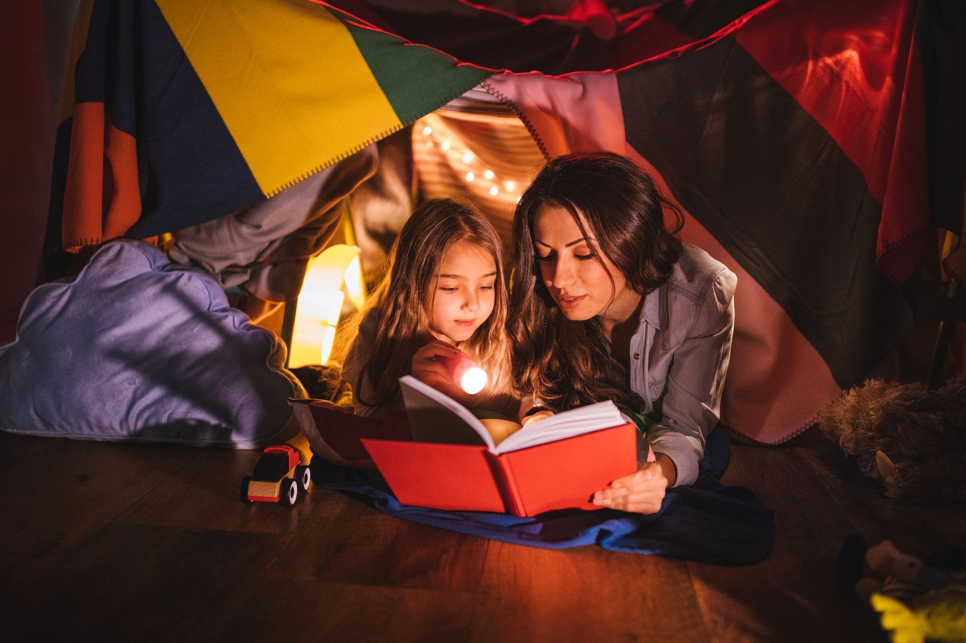 Woman and child reading a book inside a blanket fort, lit by a flashlight.