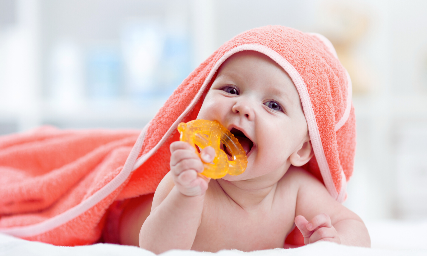 Baby wrapped in pink towel, biting an orange teething ring, smiling.