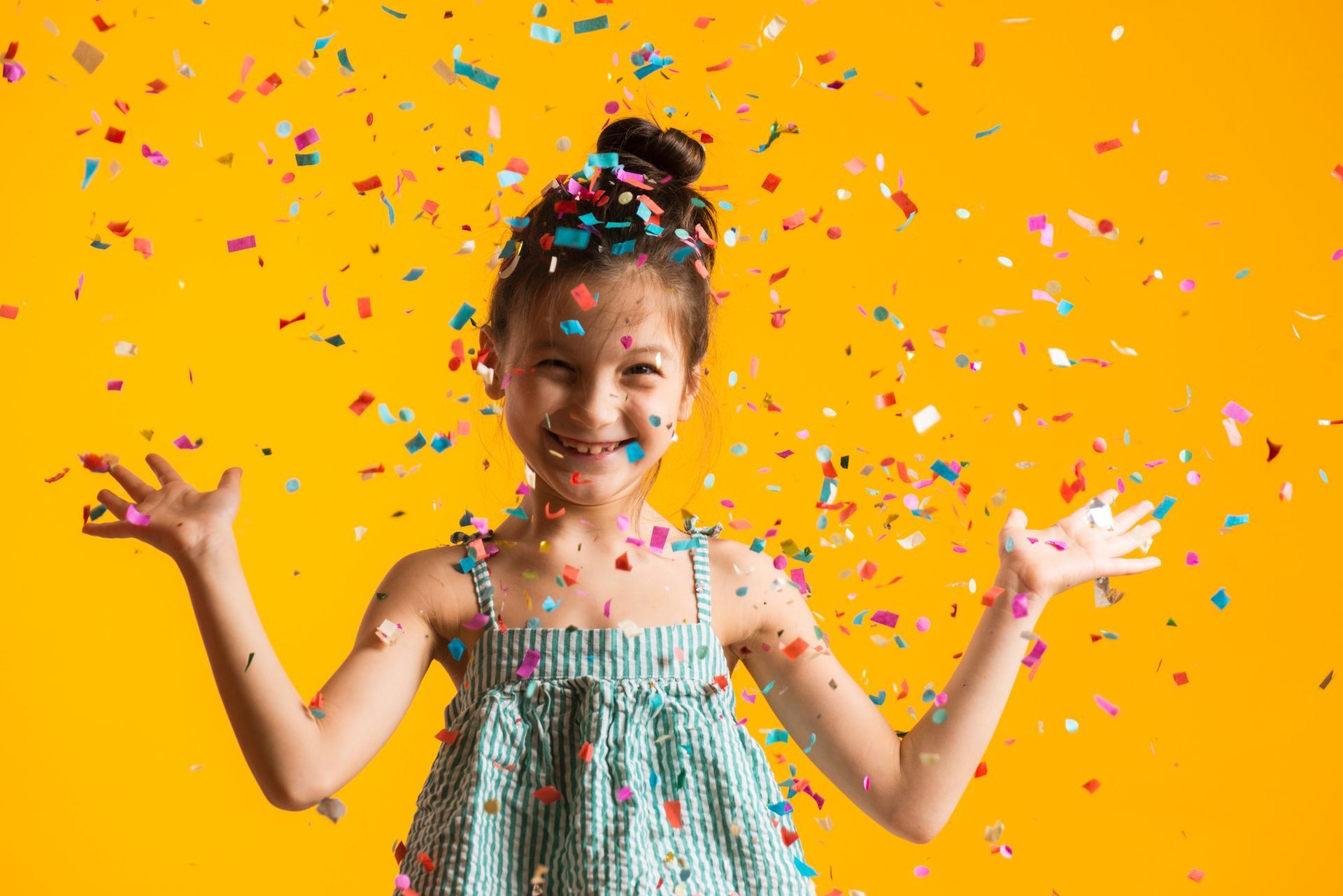 Girl with confetti in the air, smiling and raising arms against a yellow background.