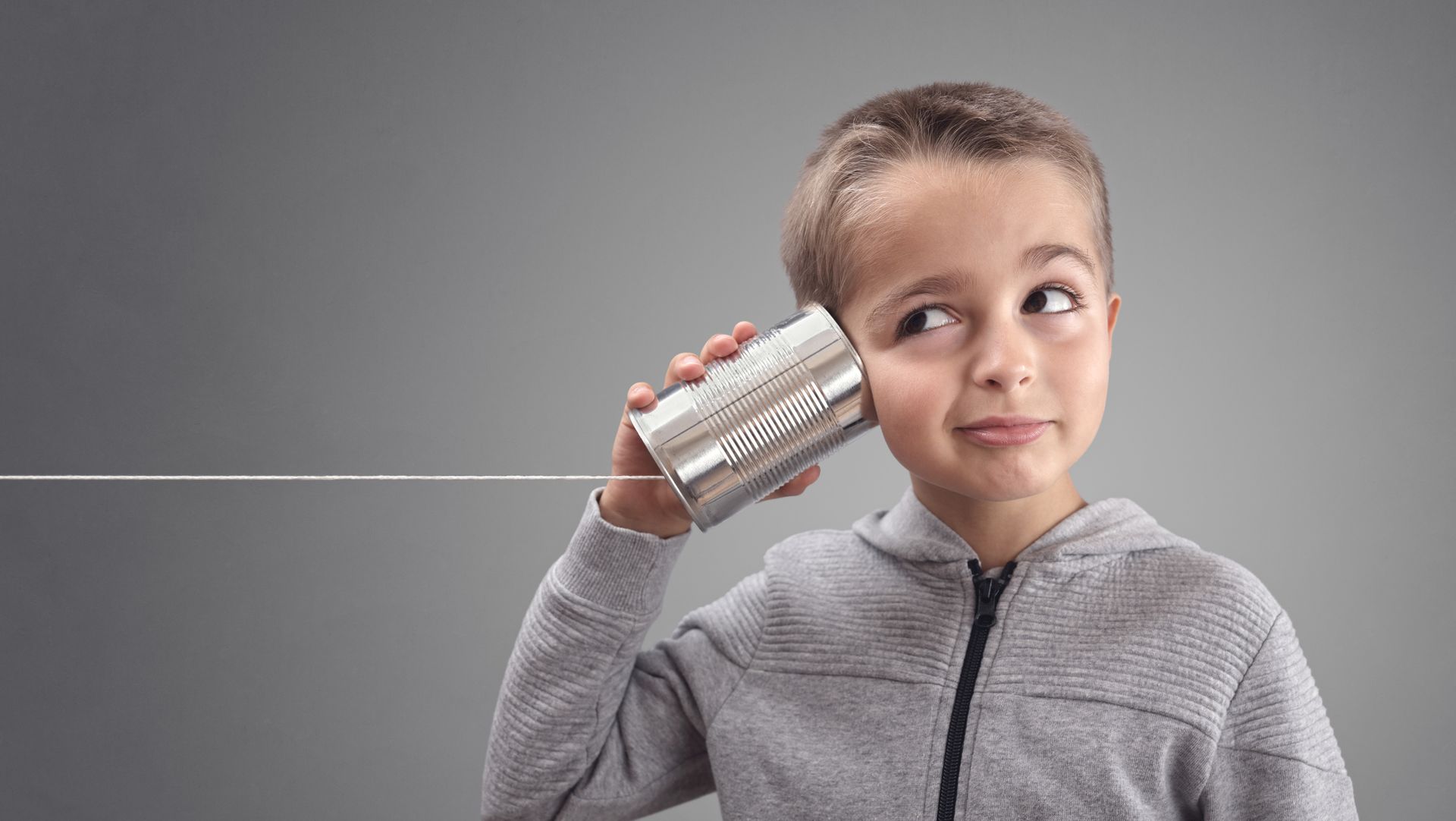 Boy holding a tin can phone to his ear, looking upwards with a curious expression. Grey background.