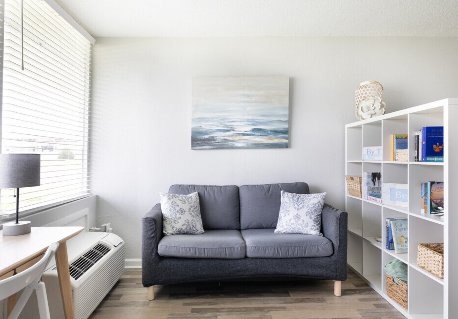 A living room with a couch , desk , shelves and a painting on the wall.