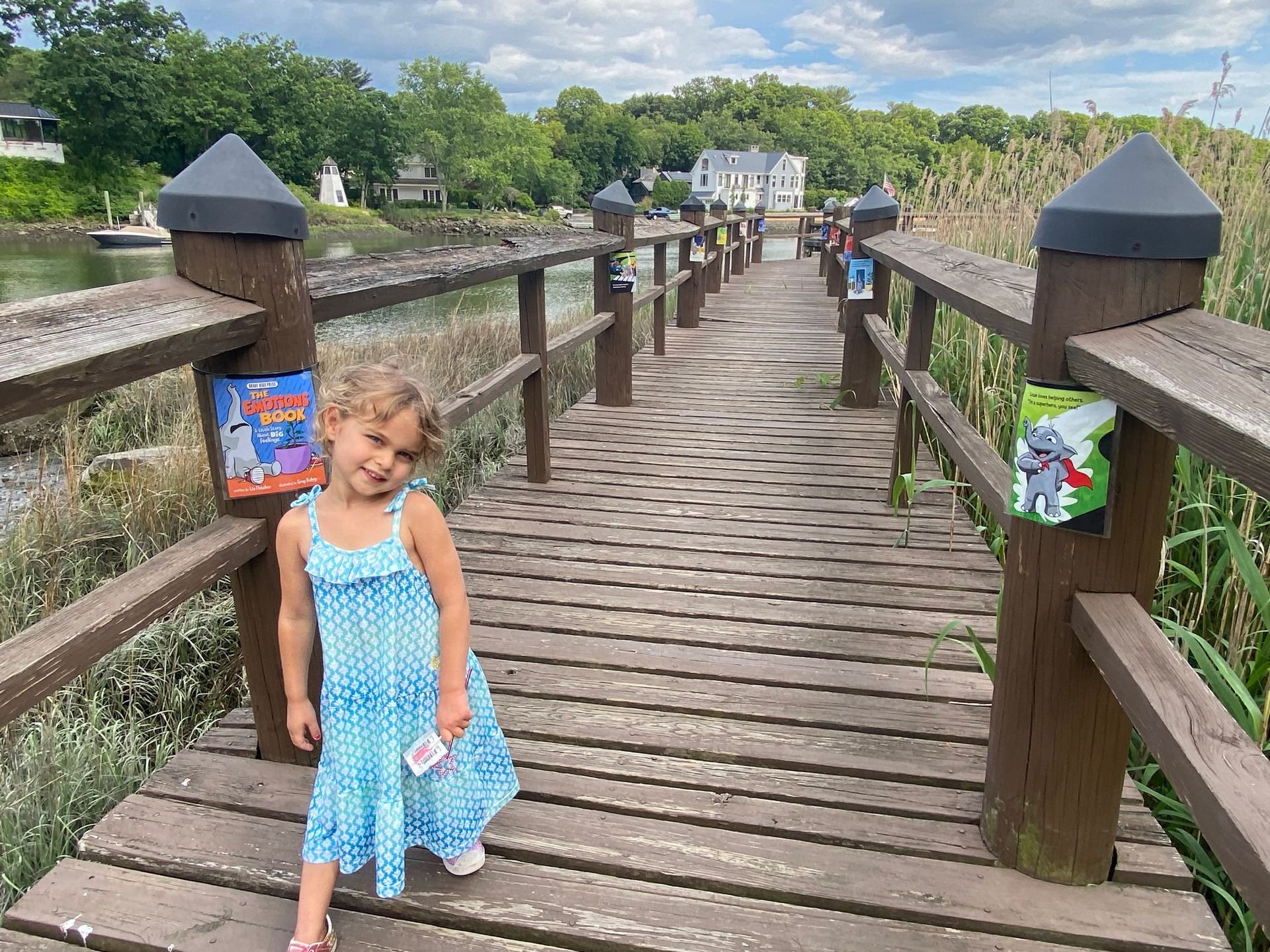 A little girl in a blue dress is standing on a wooden bridge.