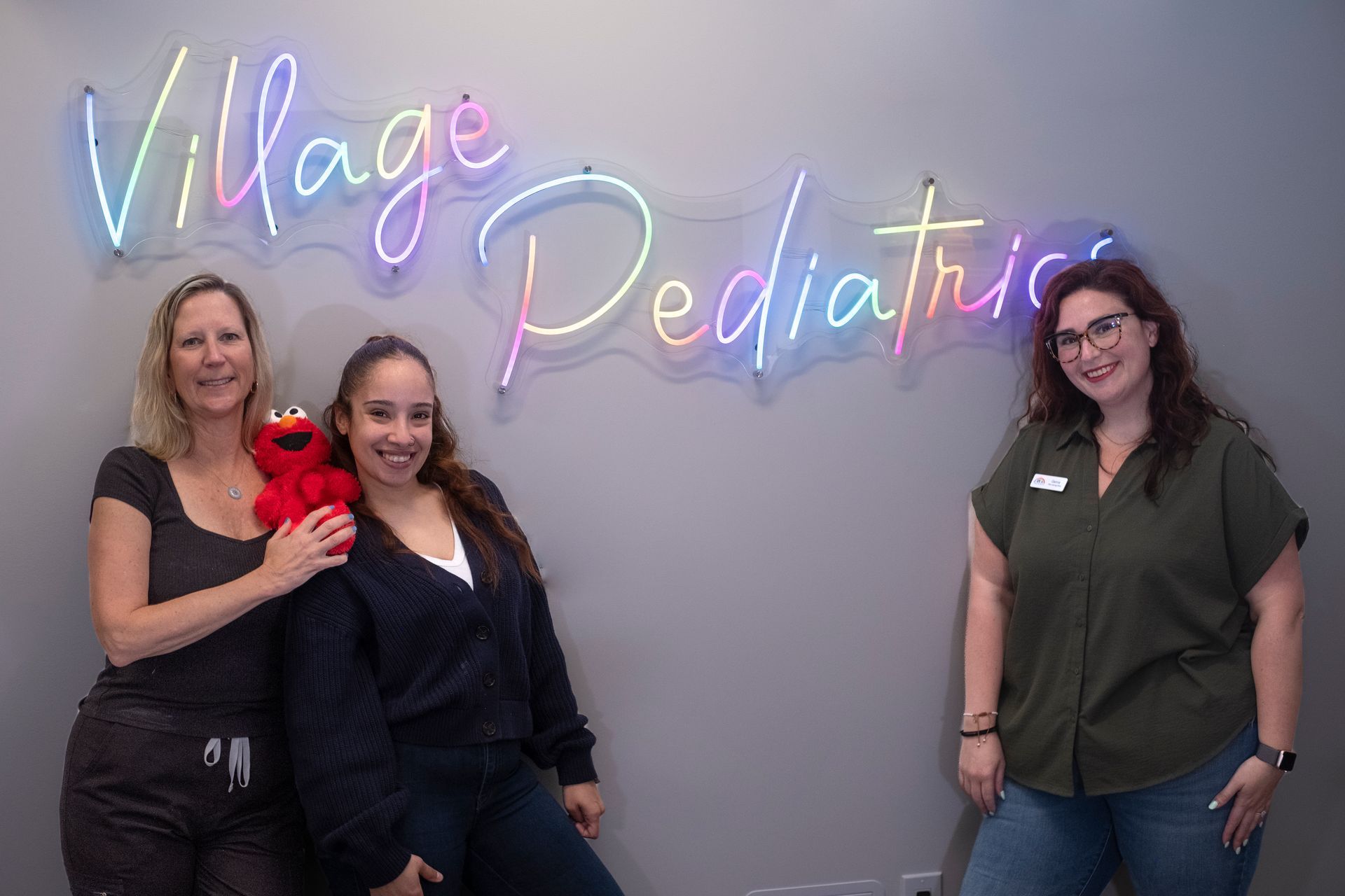 Three women are posing for a picture in front of a neon sign that says village pediatrics.