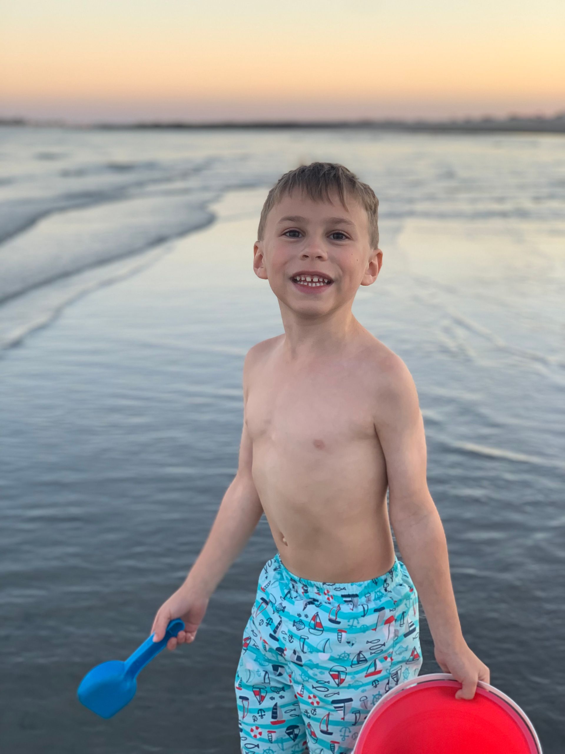 A young boy is standing on the beach holding a red ball and a blue shovel.