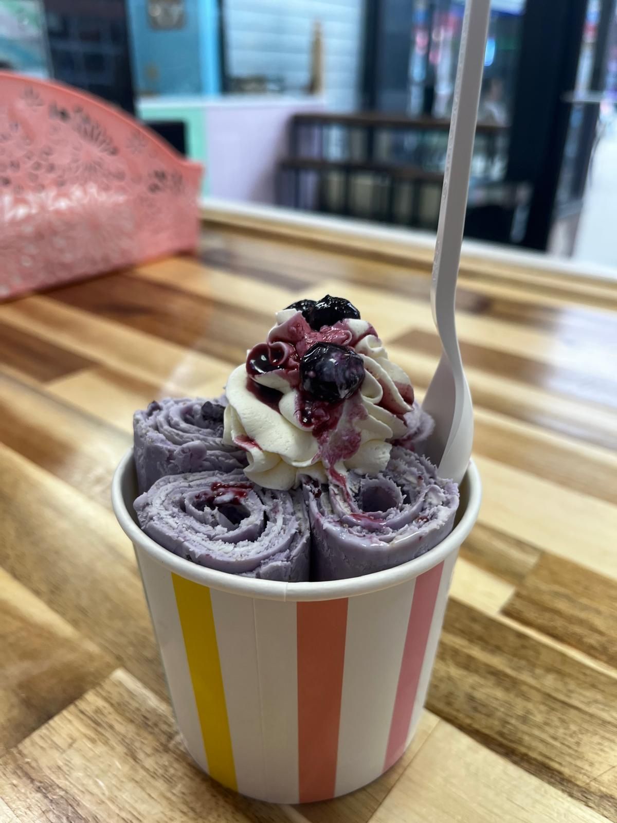 Rolled ice cream in a striped cup, topped with whipped cream and berries, with a spoon.