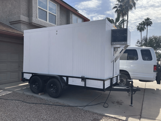 White enclosed dual-axle cargo trailer parked in a driveway beside a white van and house garage.