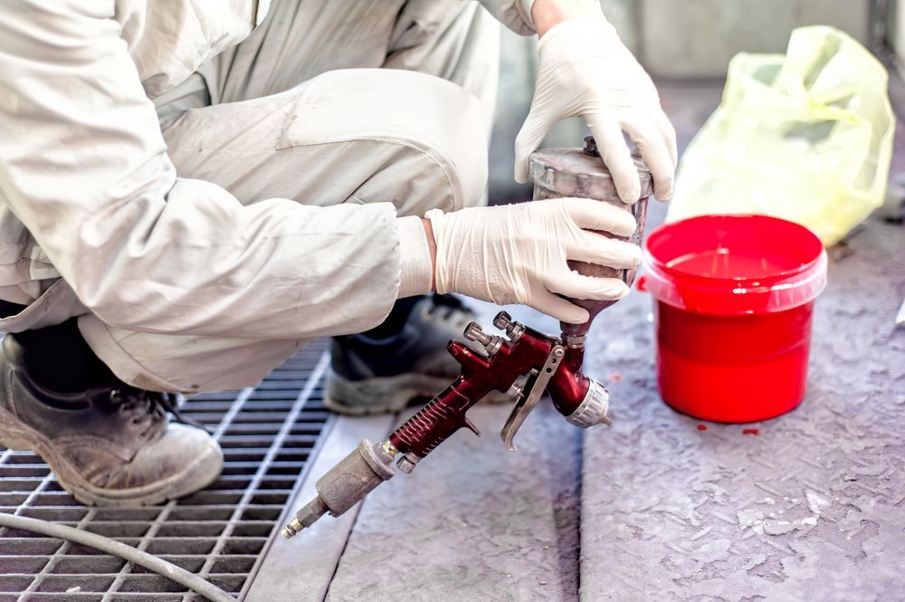 Persona con equipo de protección se prepara para pintar con aerosol; hay un balde de pintura roja cerca.