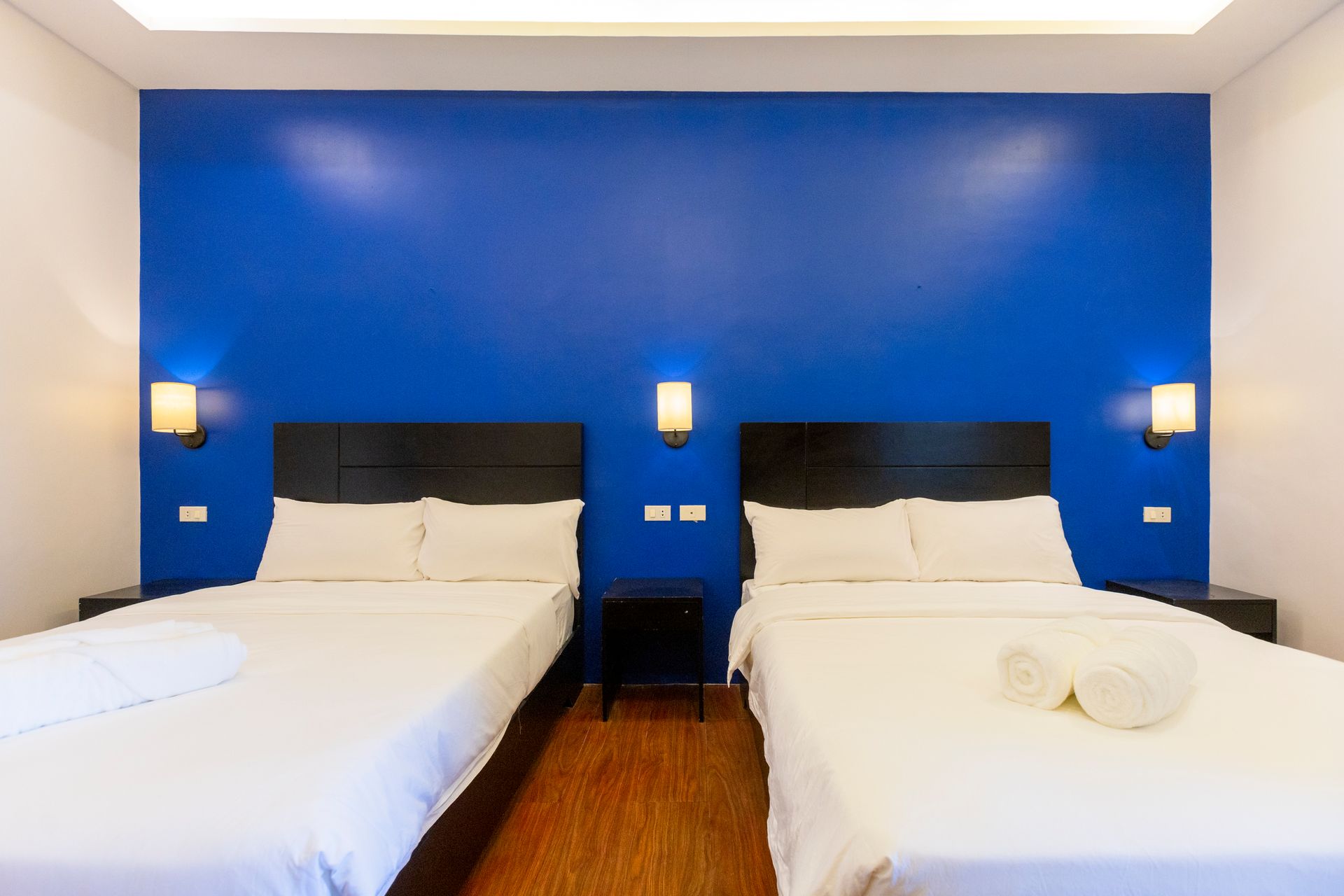 Two beds with white bedding, against a blue wall with sconce lights and dark headboards in a room.