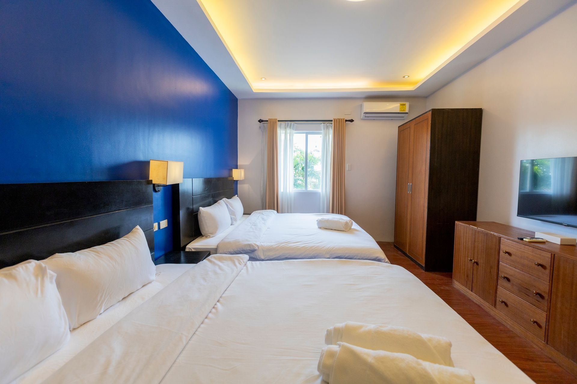 Hotel room with three beds, blue wall, wooden furniture, and window with curtains.