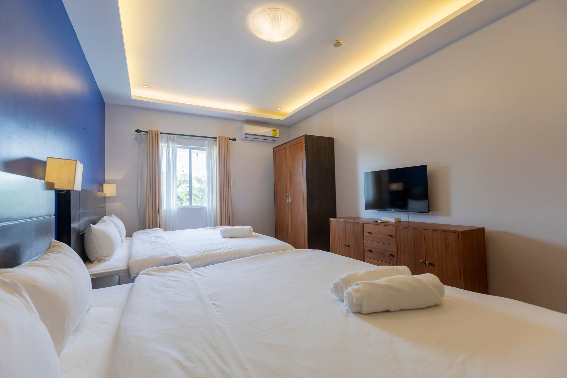 Bedroom with two white beds, a TV, and a wooden wardrobe. Blue and white walls, natural light.