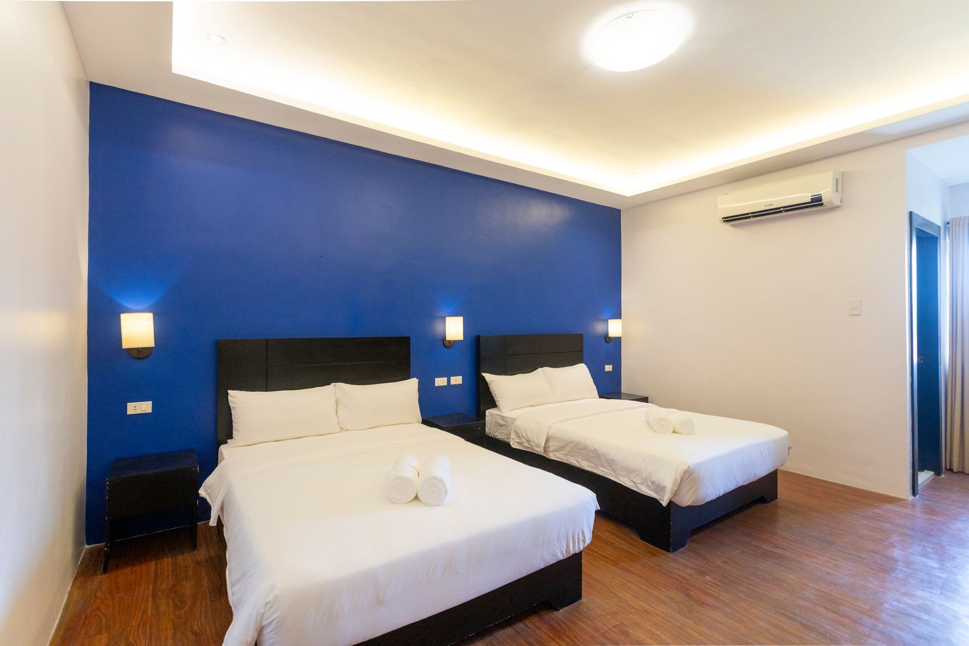 Hotel room with two beds, blue accent wall, neutral walls, wood floor, and overhead lighting.