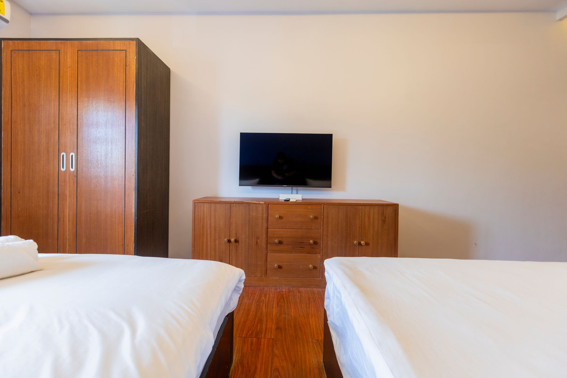 Bedroom with two beds, wooden wardrobe, TV, and dresser. White bedding, beige walls, and wood floor.