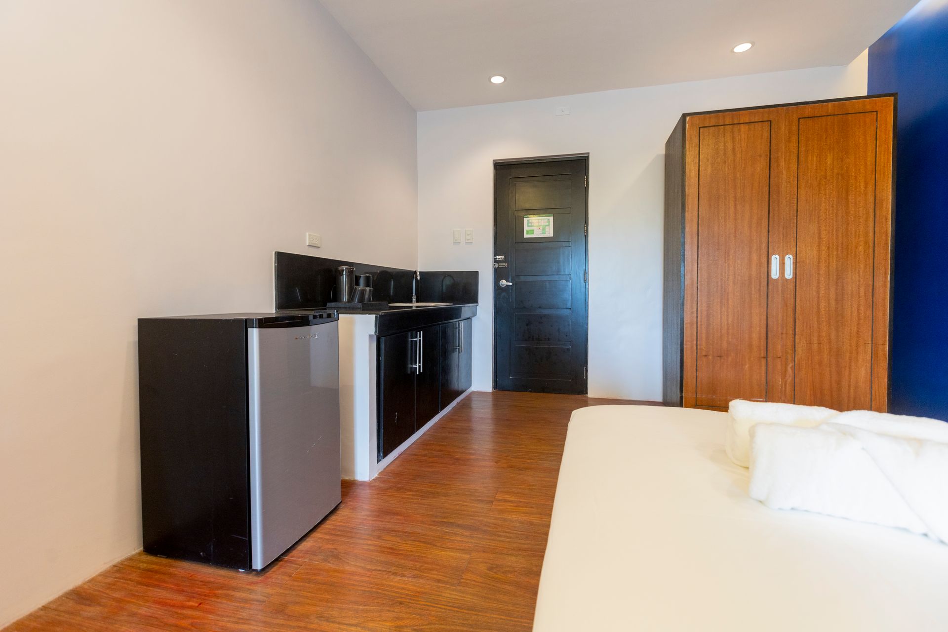Small studio apartment with kitchenette, fridge, closet, bed, and black door.
