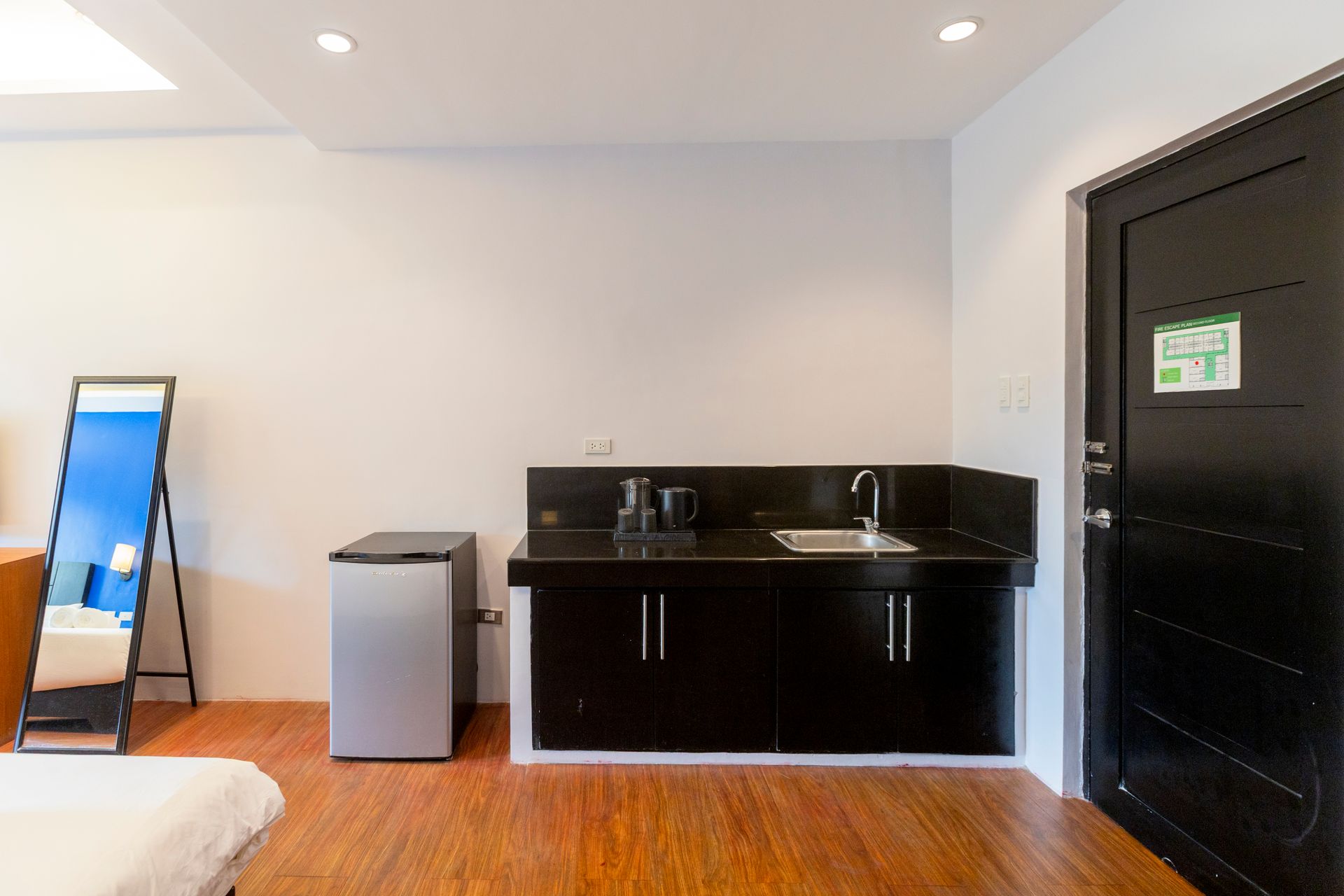 Room interior: kitchenette with black cabinets, sink, mini-fridge, and mirror. White walls, wood floor.