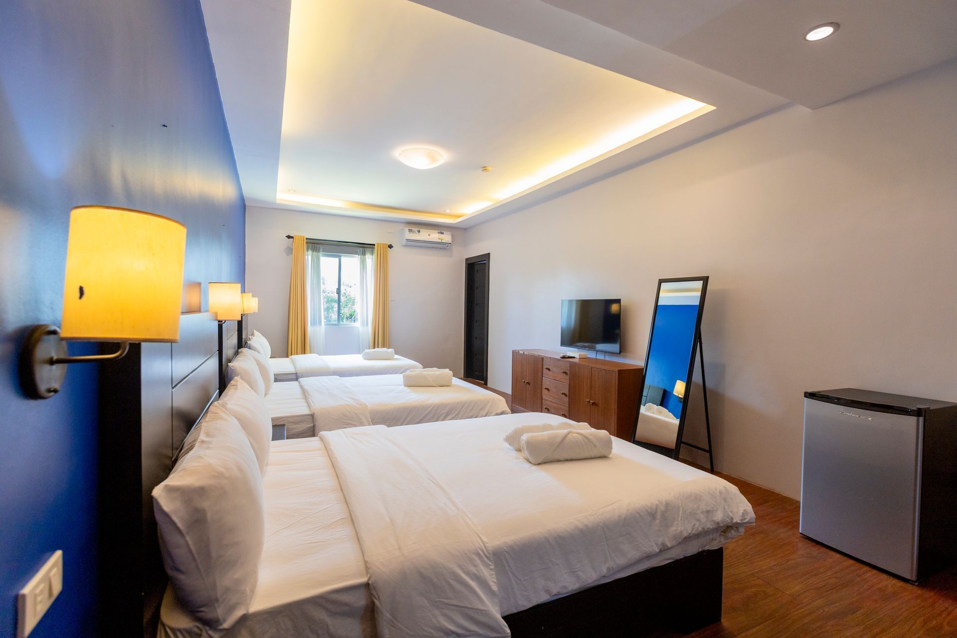 Hotel room with three beds, TV, mirror, and small refrigerator. Walls are blue and white.