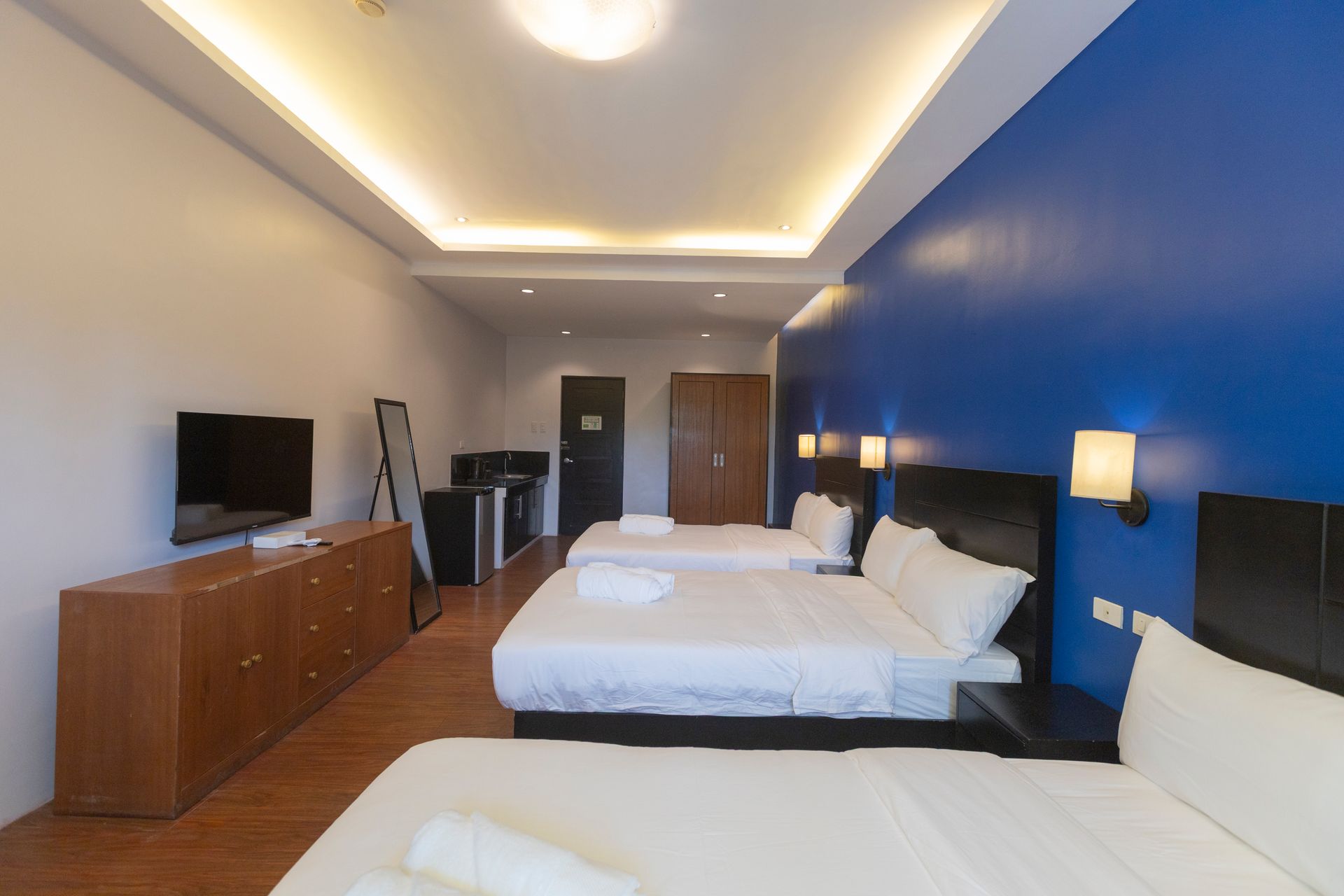 Hotel room with three beds, blue accent wall, TV, and wooden furniture.