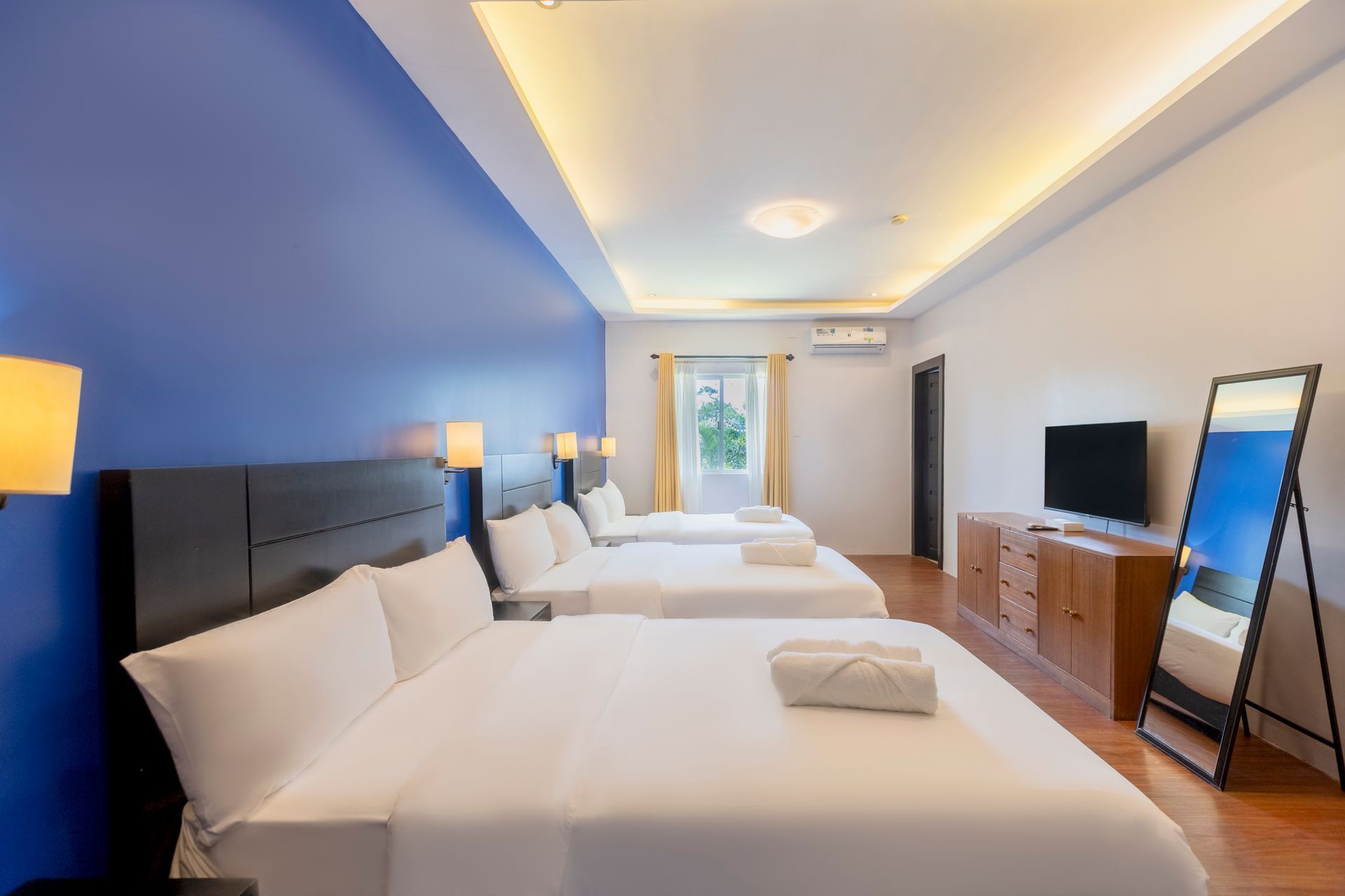 A hotel room with three beds, blue wall, wooden furniture, and a full-length mirror.