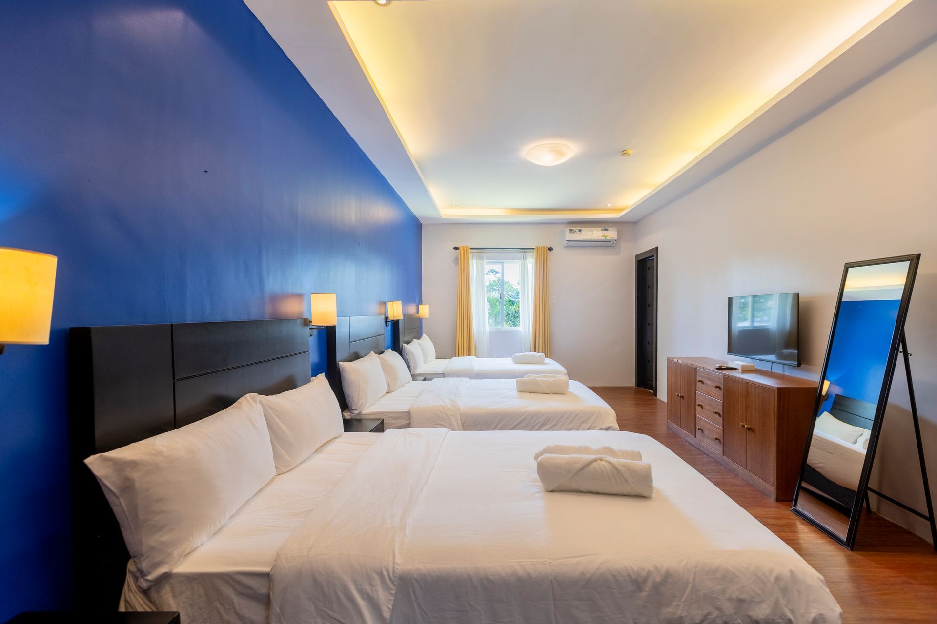 Hotel room with three beds, a mirror, and a TV, blue accent wall.