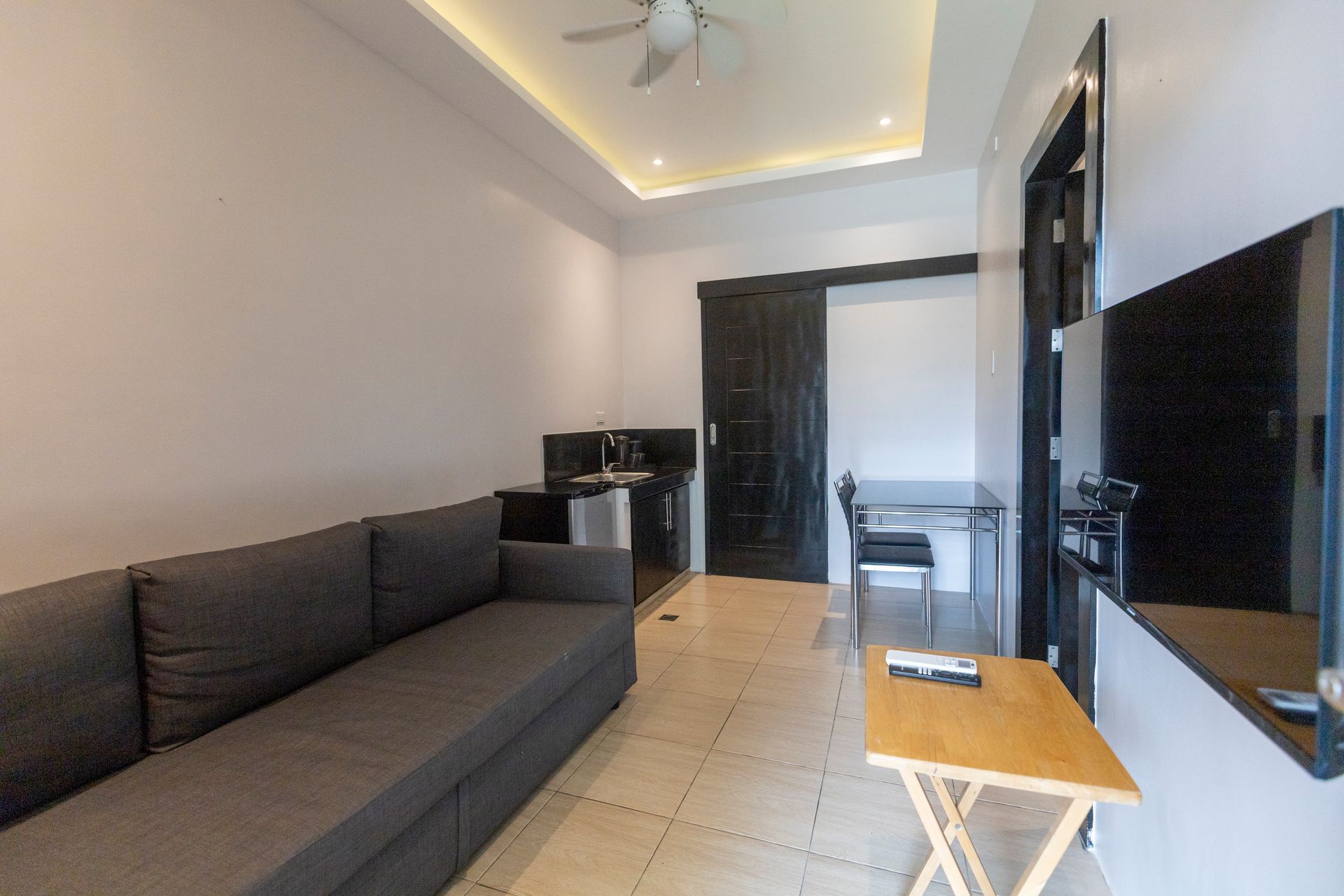Small apartment interior with gray sofa, kitchen, table, and mounted TV.