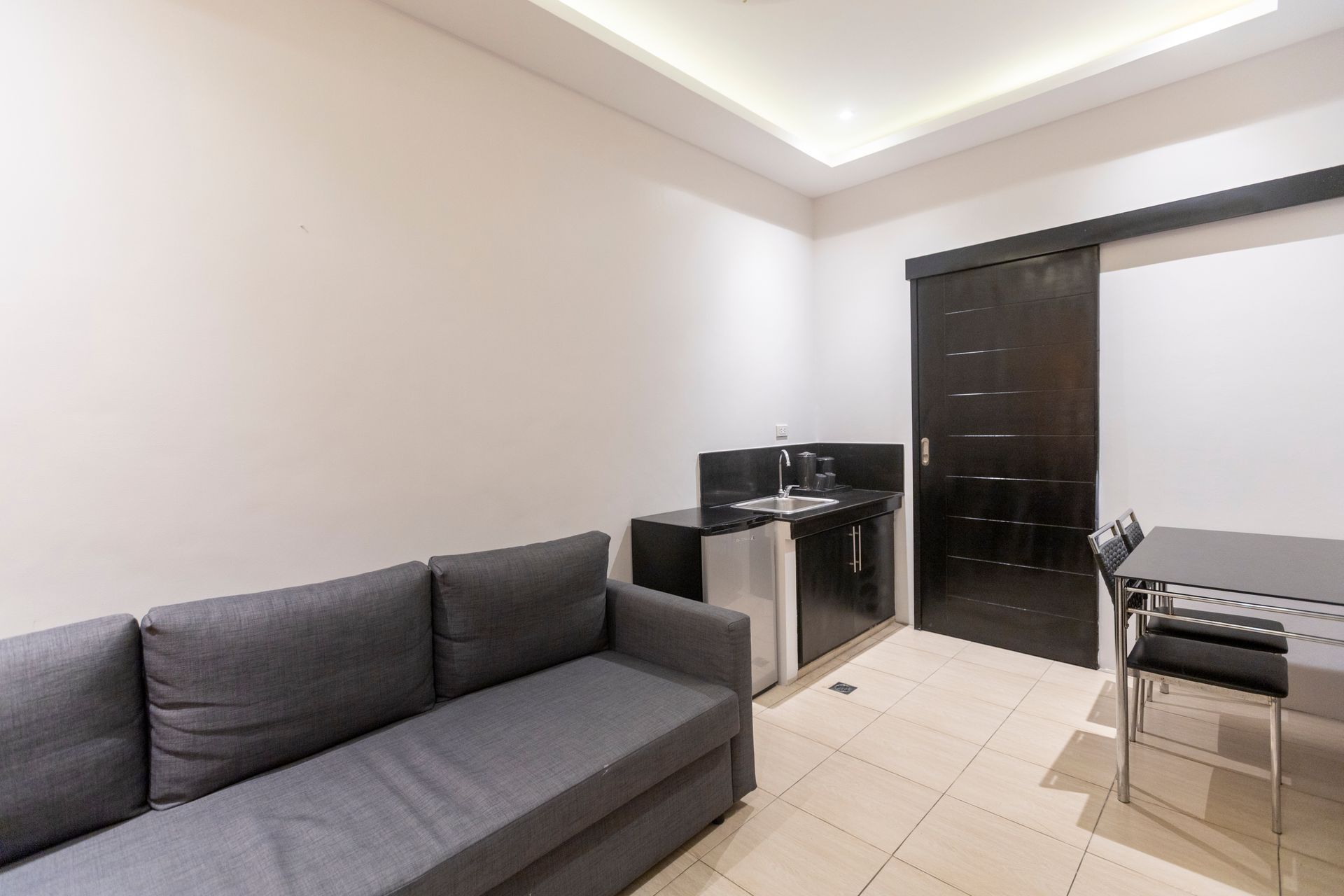 A small apartment with a gray sofa, kitchenette, dark wood door, and dining table. White walls, tile floor.