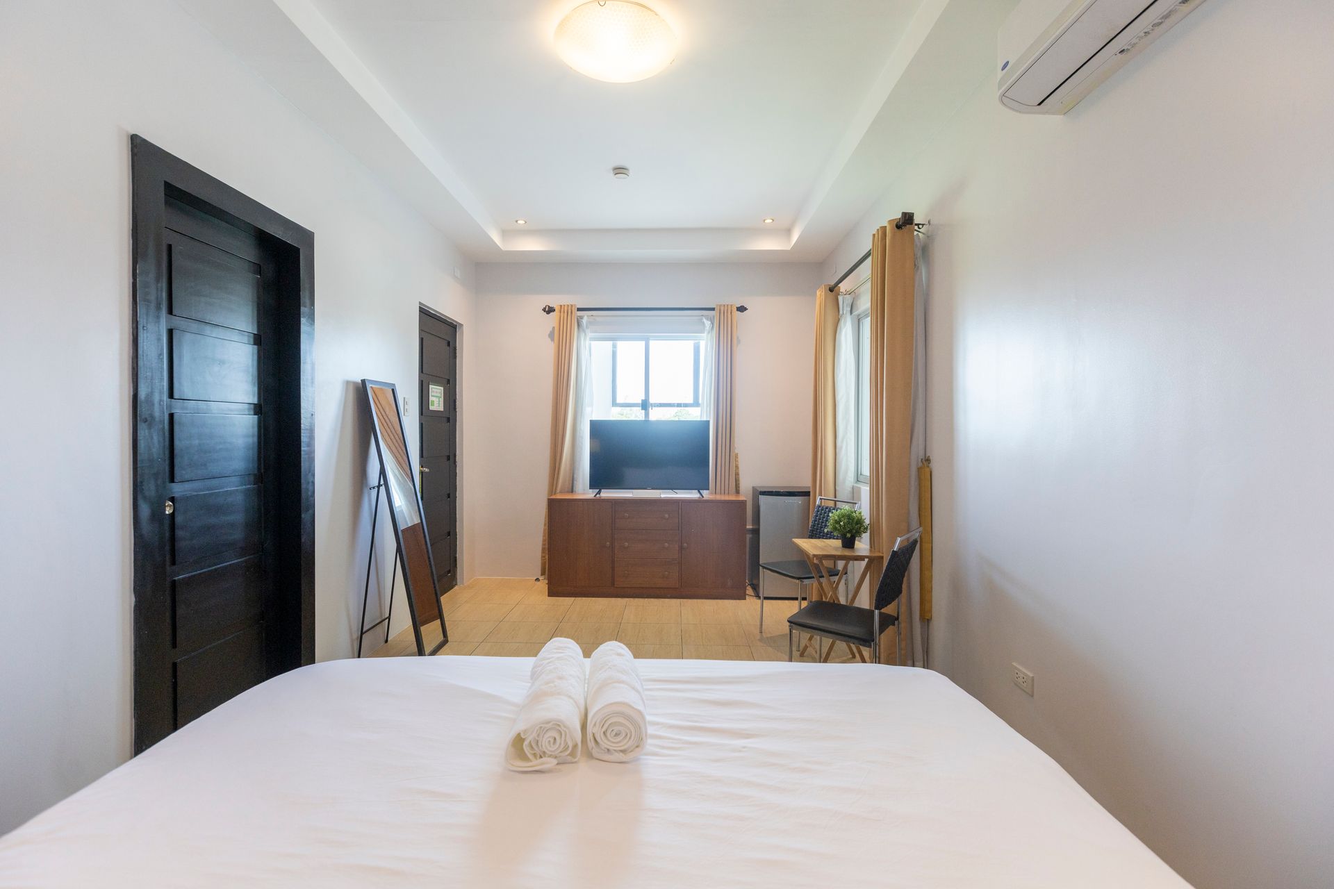 Bedroom with a bed, TV, and side table. White walls, wooden furniture, and folded towels.