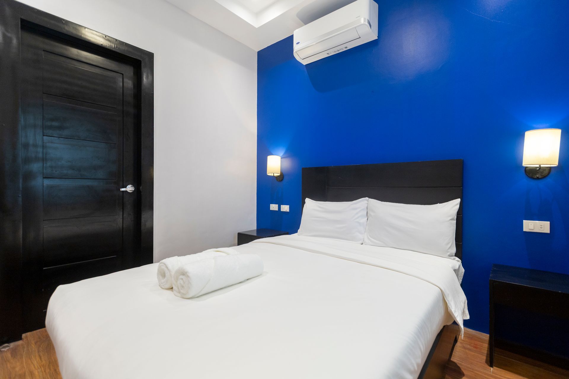 Bedroom with a white bed, black door, and blue wall, with air conditioning and two wall lights.
