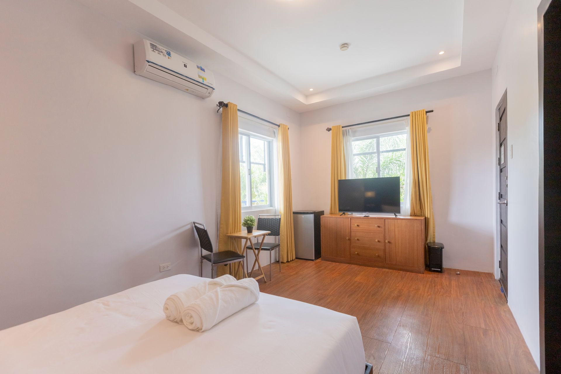 Bedroom with white walls, wood floor, two windows, TV, and air conditioner. A rolled towel lies on a bed.