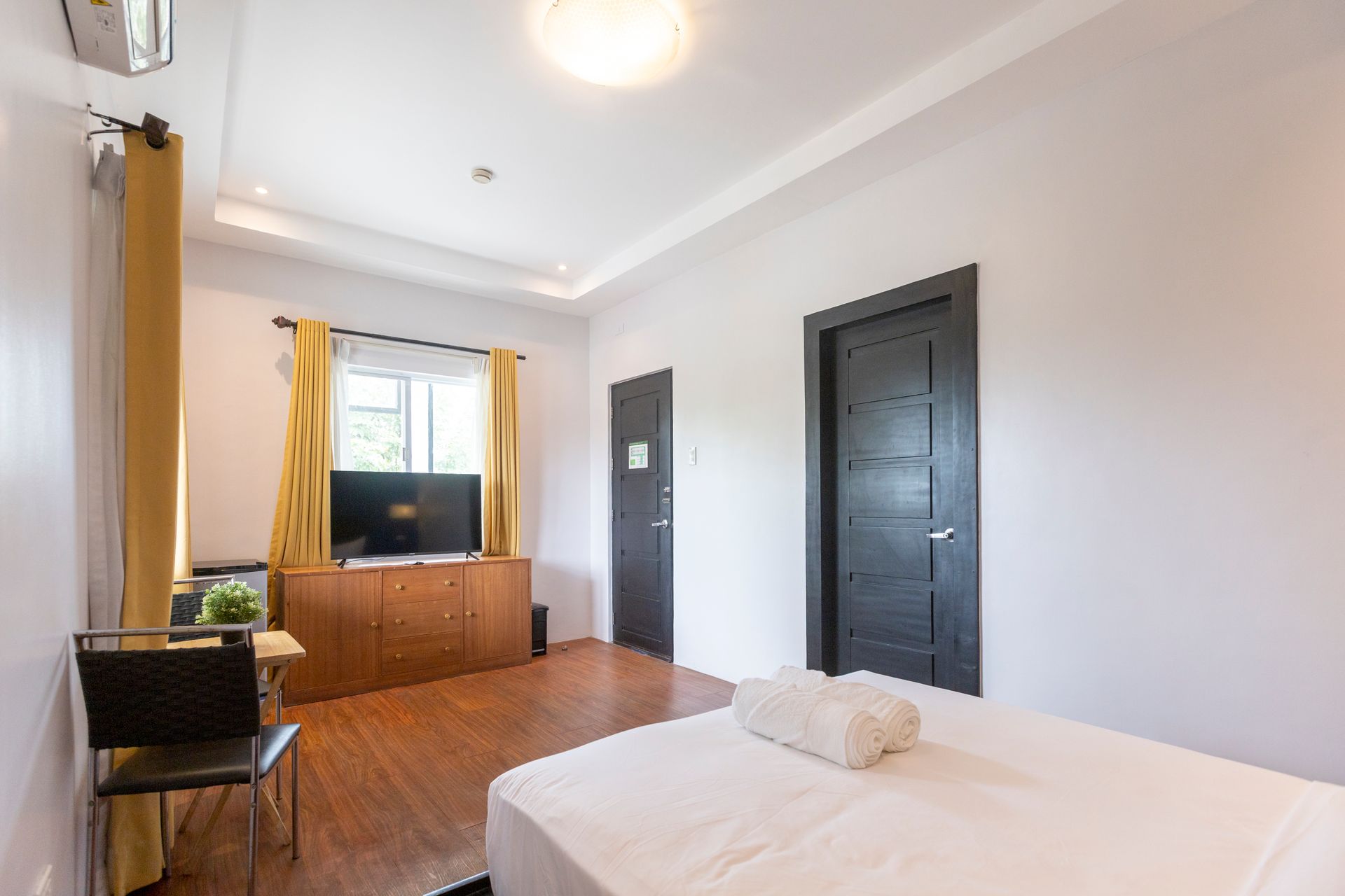 Bedroom with a bed, TV, dark doors, and wooden floor. Yellow curtains and neutral walls.