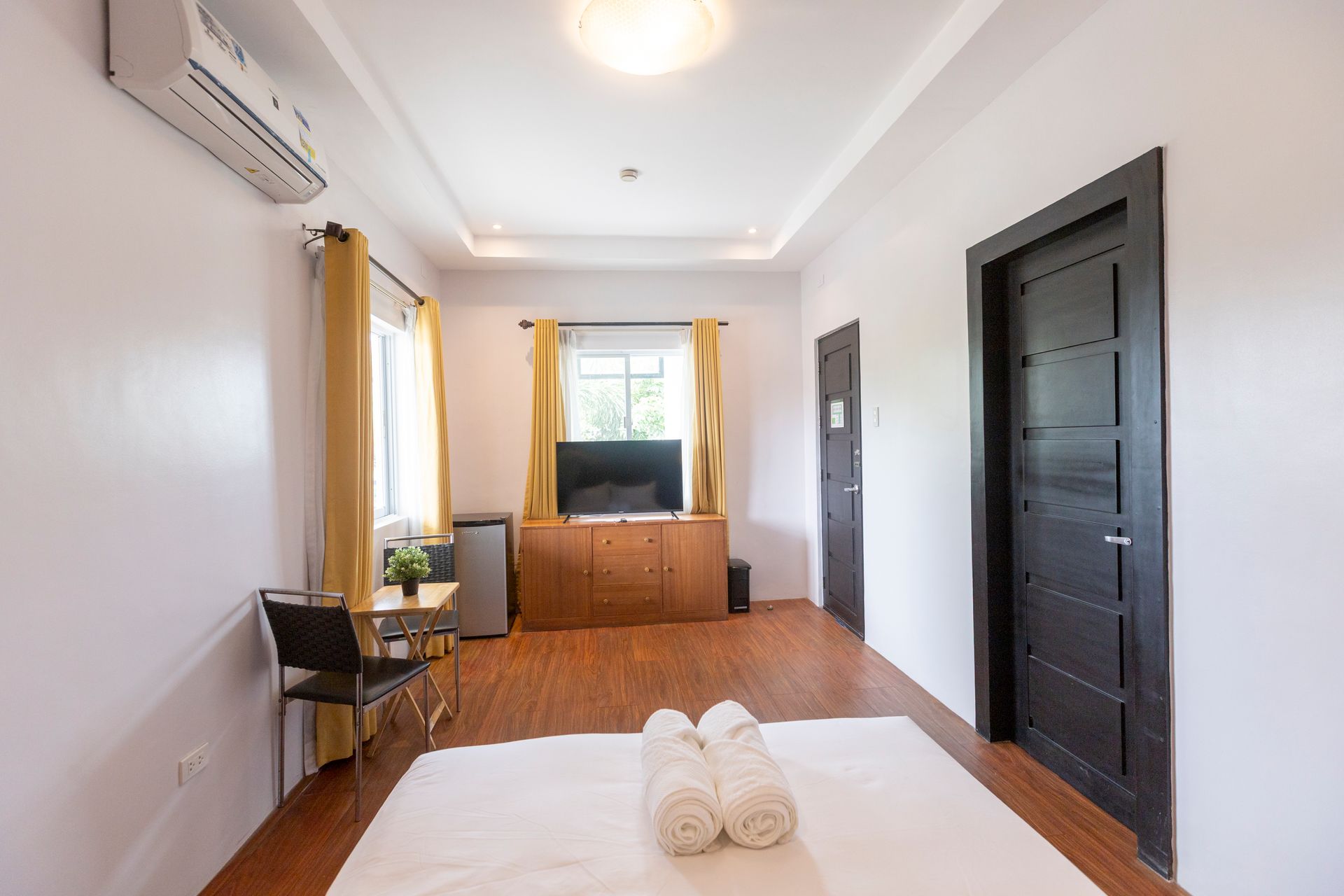 Hotel room with bed, TV, dark door, window with yellow curtains, and air conditioning.