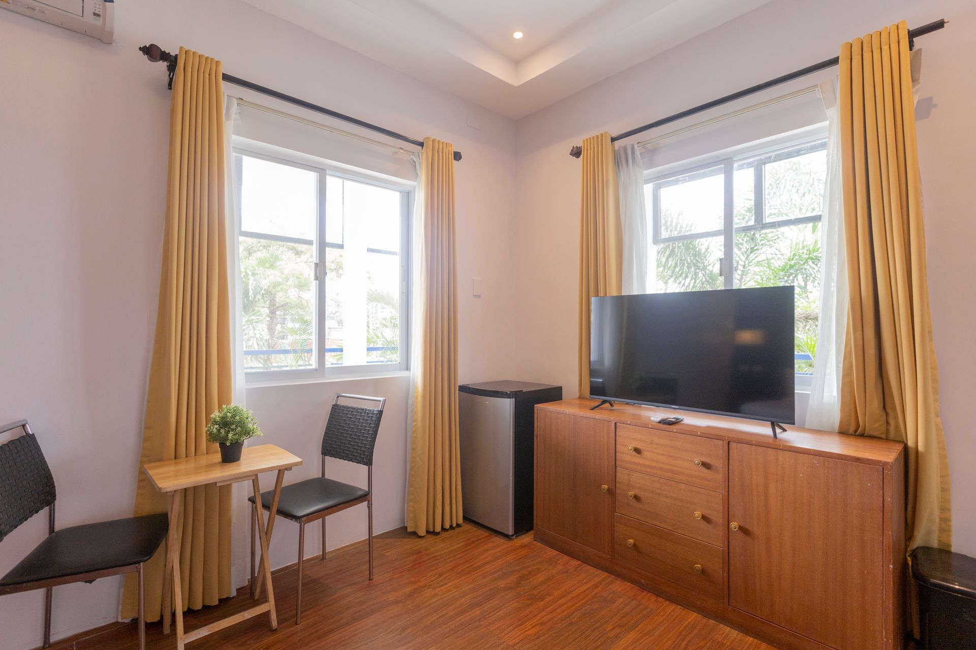 A bedroom with two windows, TV on wooden cabinet, small table with plant, two chairs, and yellow curtains.