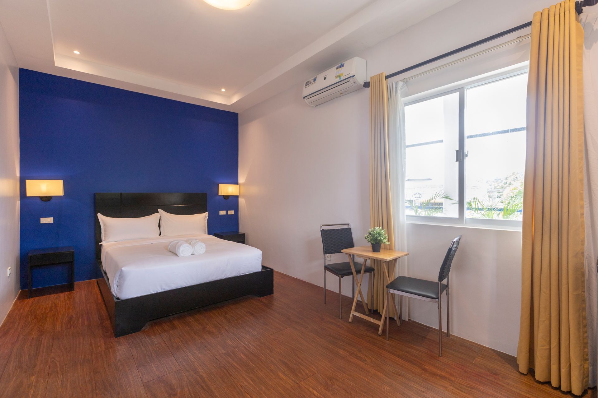Hotel bedroom with bed, blue accent wall, window, two chairs, and a small table.