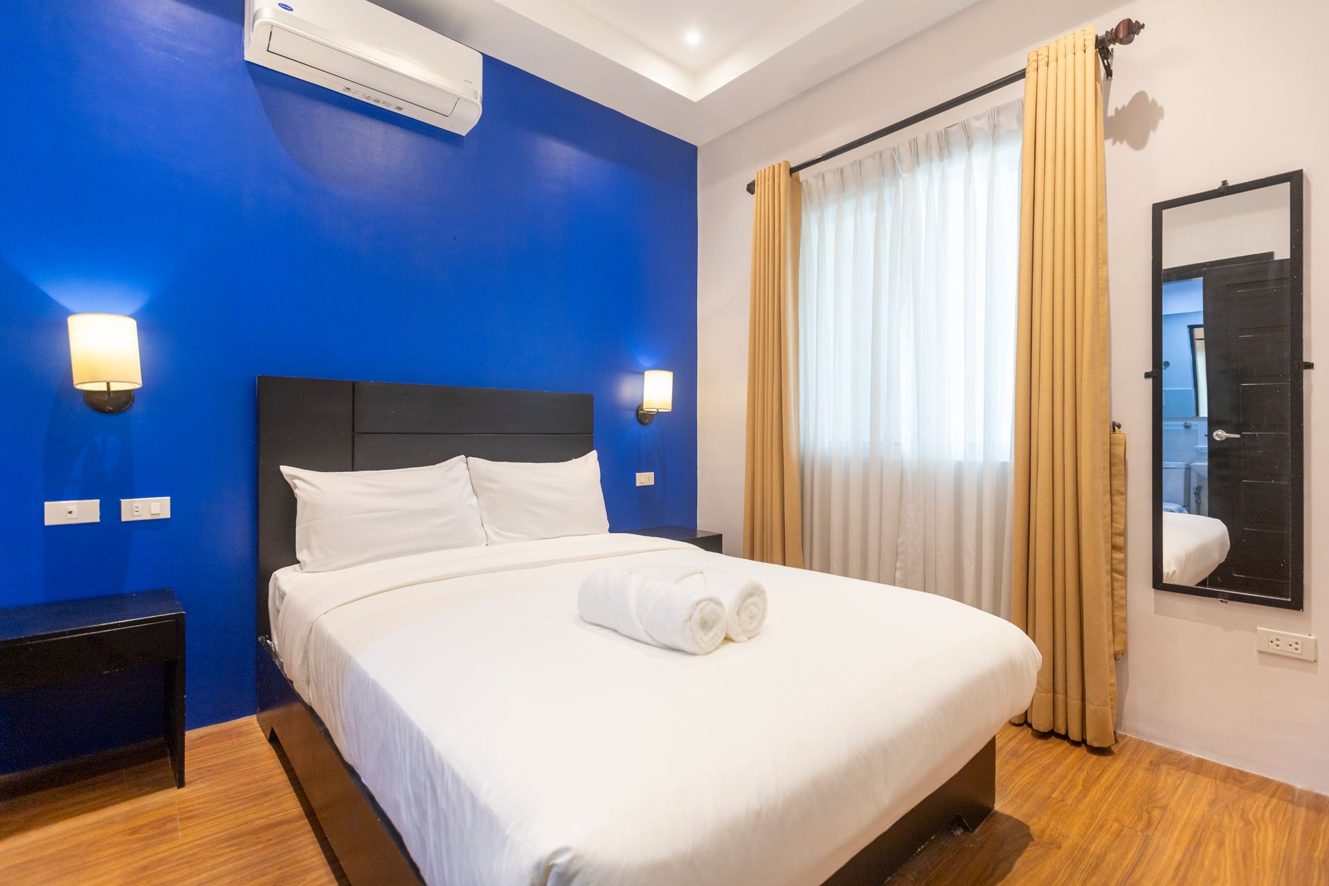 Bedroom with a blue wall, white bed, golden curtains, and a black-framed mirror.