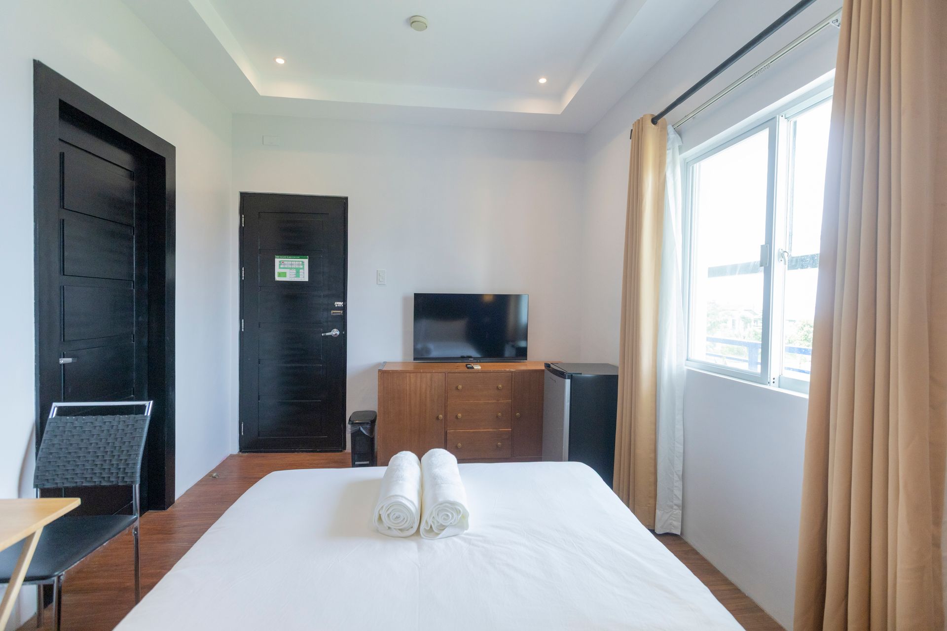 Hotel room with bed, TV, window, black door, and small fridge.
