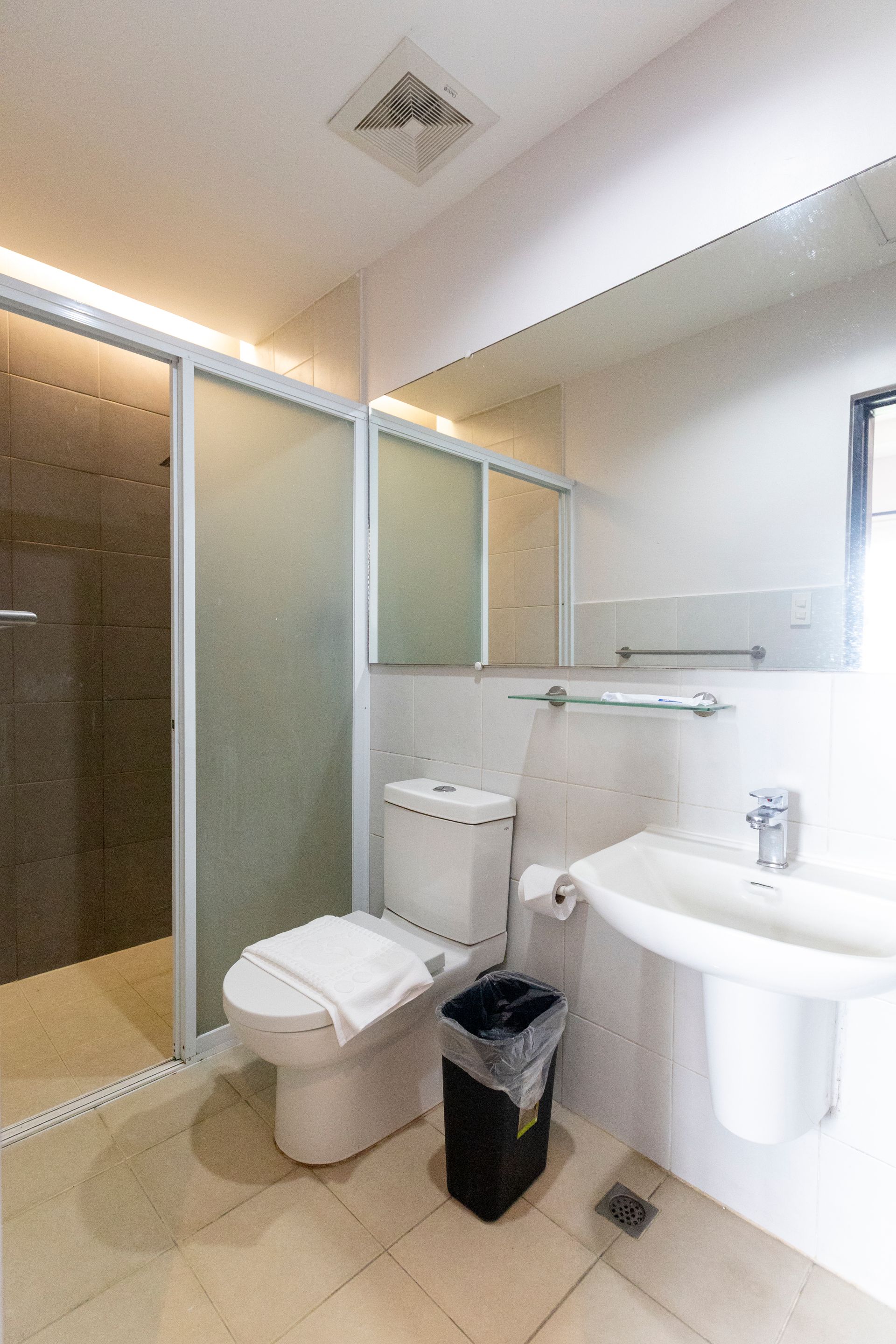 Bathroom with toilet, sink, shower, and a black trash can. Walls are white and floors are beige.